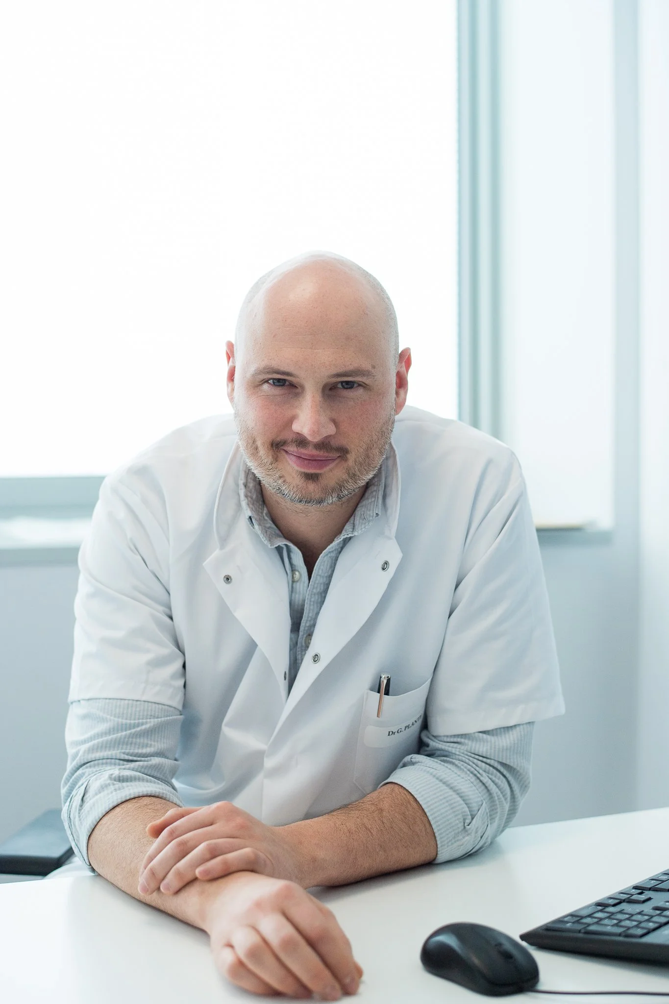 Un homme chauve portant une blouse blanche assis à un bureau avec un ordinateur dans un environnement lumineux.