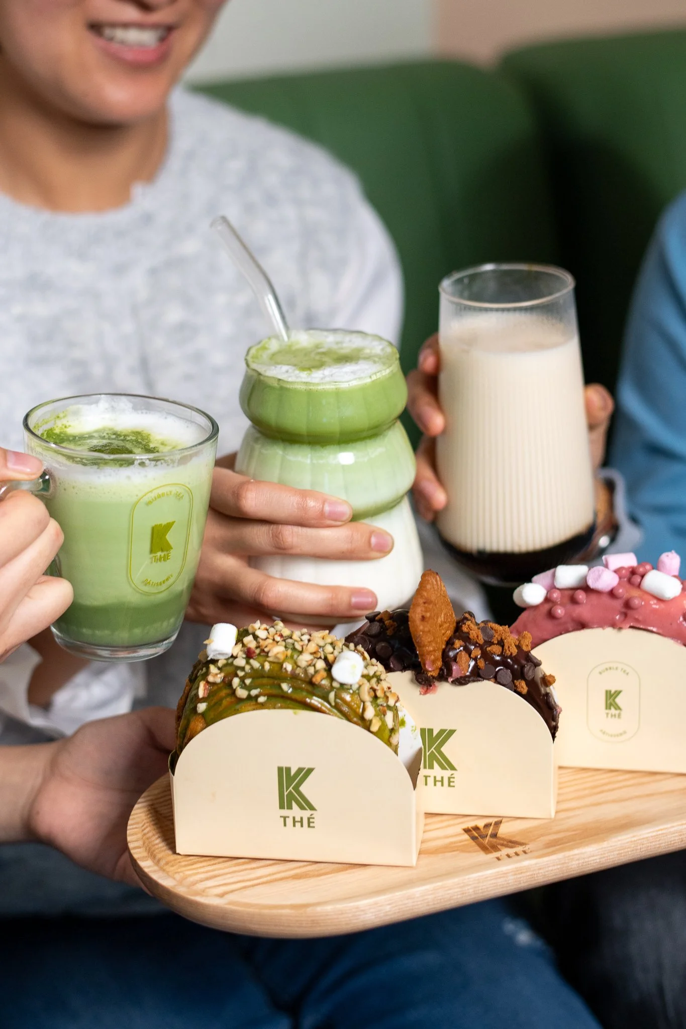 Deux personnes tenant des boissons et desserts, avec des desserts variés comme un gâteau garni de topping, et deux boissons, l'une verte et l'autre blanche, sur une table en bois.