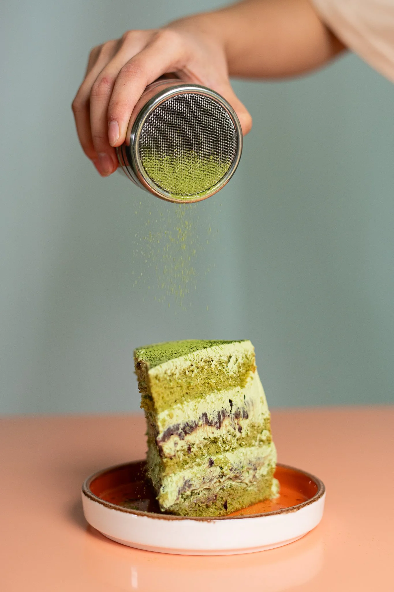 Une main saupoudre de la poudre de matcha sur un gâteau vert à plusieurs couches, placé dans une petite assiette blanche.