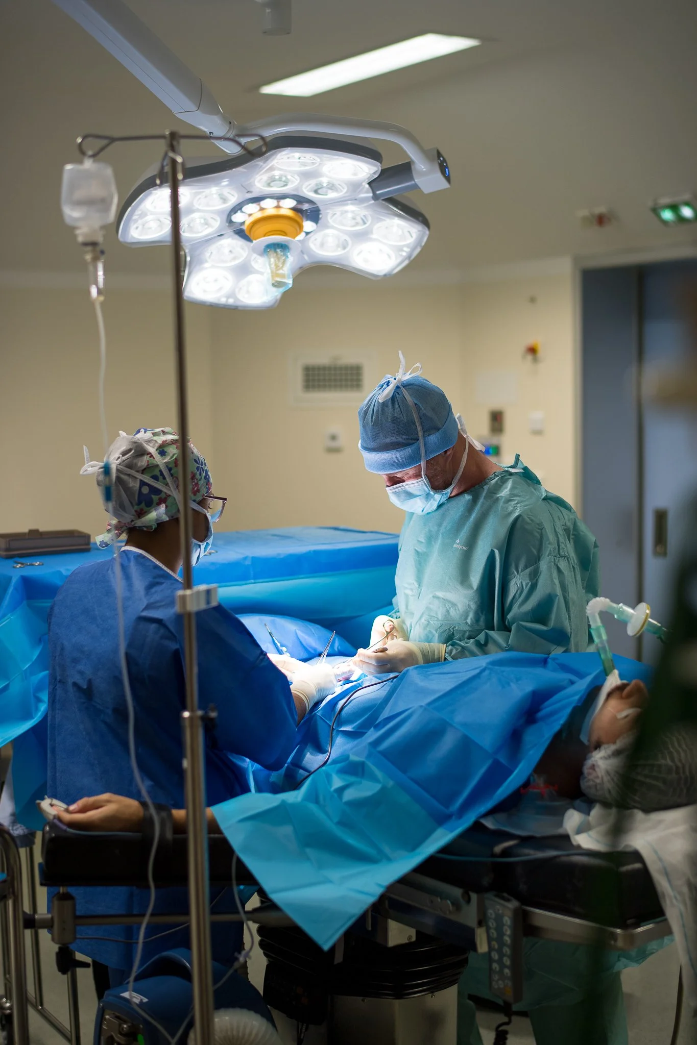 Chirurgiens en train d'opérer dans une salle d'opération, portant des blouses, des masques et des couvre-cheveux. Des équipements médicaux et un plafond lumineux sont visibles.