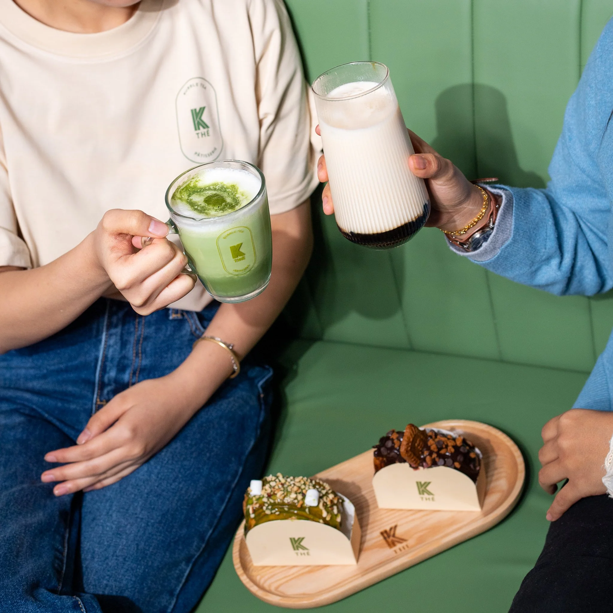 Deux personnes qui tiennent des verres de boissons colorées, l'une avec une boisson verte mousseuse et l'autre avec une boisson blanche et noire. Sur une assiette en bois, il y a deux desserts avec du chocolat et des biscuits, chaque dessert a une étiquette avec la lettre 'K' dessus.