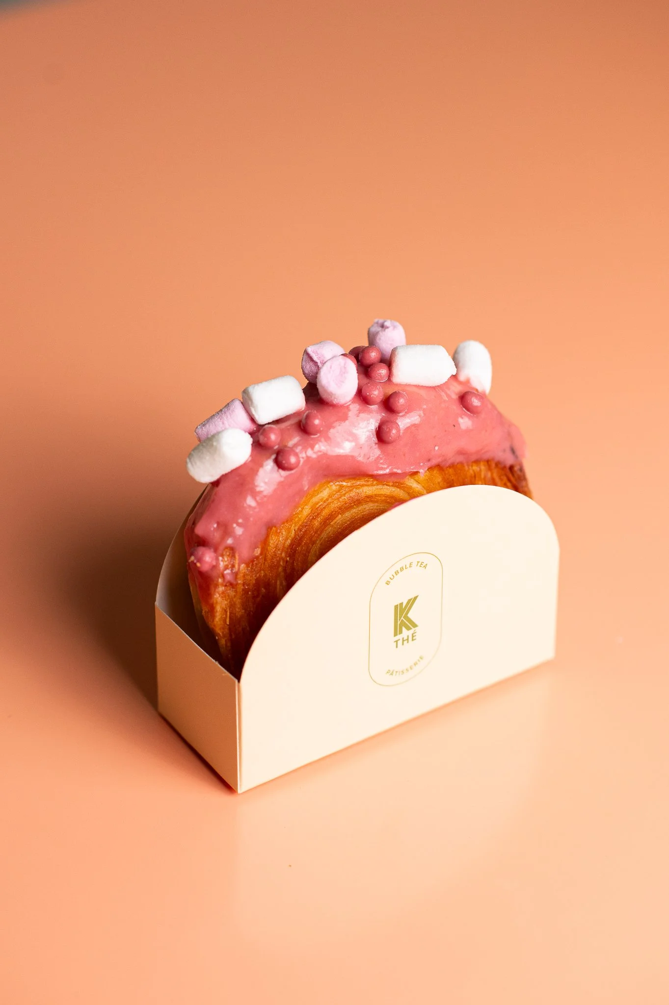Gâteau rond avec glaçage rose, décoré de petits marshmallows et perles roses, dans une boîte blanche avec logo doré, sur un fond orange clair.