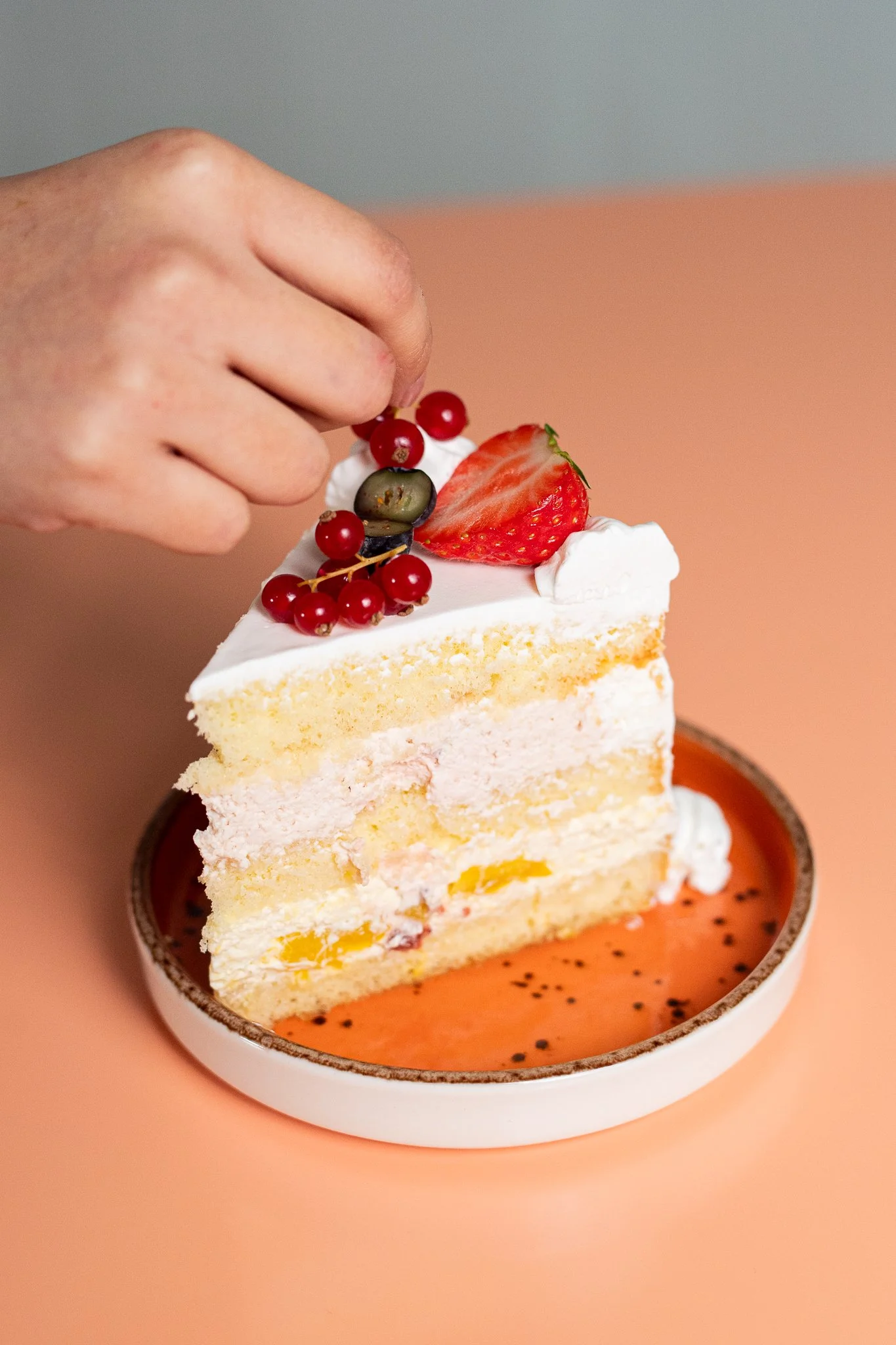 Une part de gâteau décorée de fruits rouges, une fraise tranchée, et une petite grappe de groseilles, avec une main saupoudrant ou plaçant un fruit dessus.