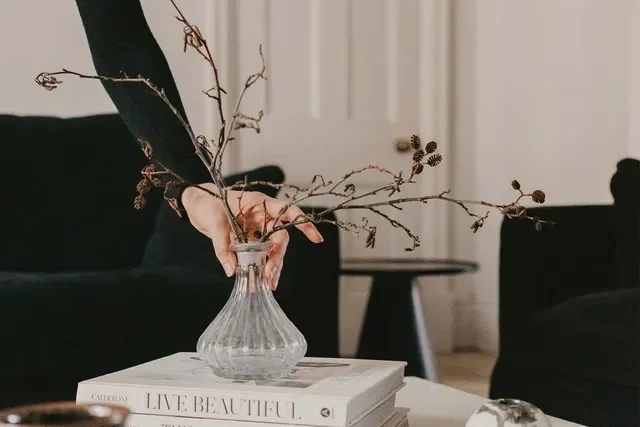 Person arranging dry branches and seed pods in a clear glass vase on top of a stack of books in a cozy living room.