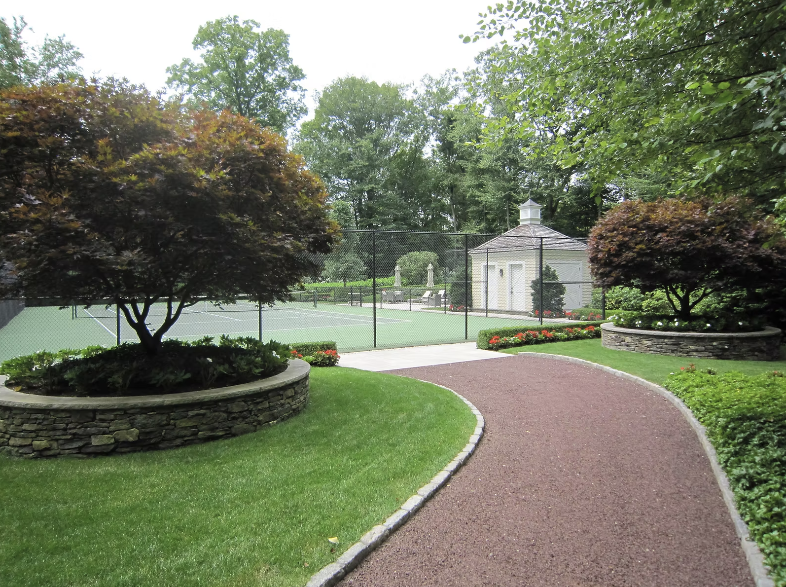 A sports court, surrounded by well-maintained lawns, colorful flowers, lush green trees, and a small building in the background.