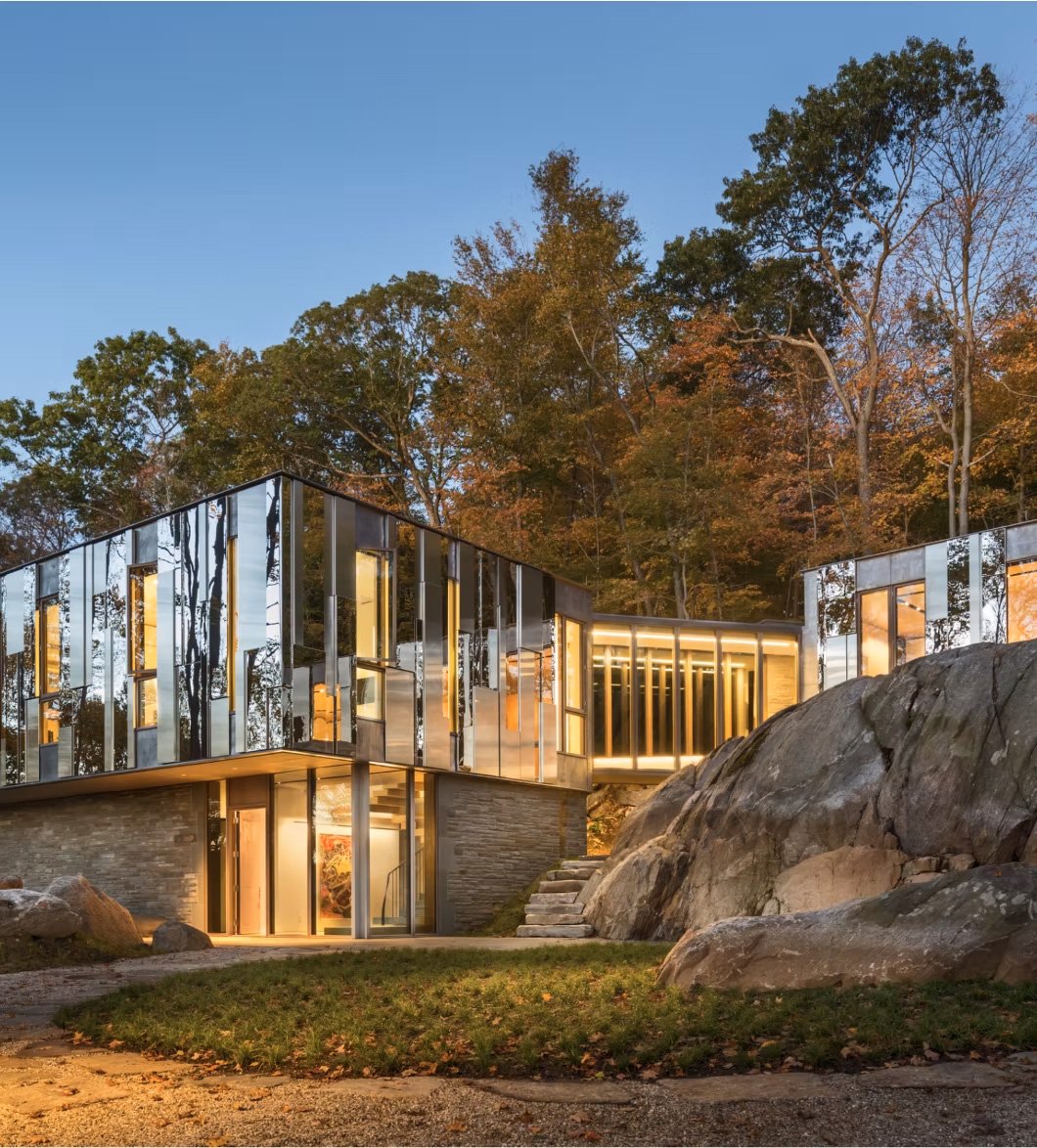 Modern house built into a rocky hillside surrounded by trees with autumn foliage, illuminated at dusk.