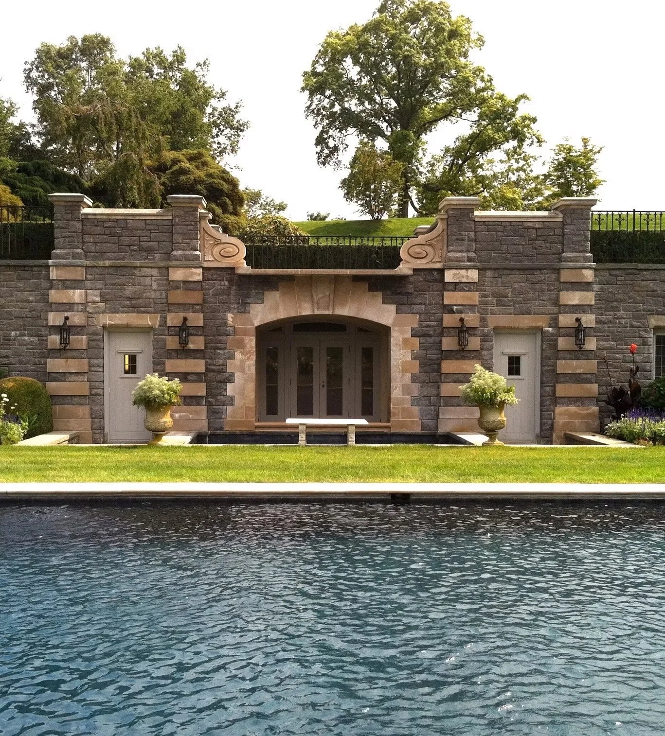 Stone house with arched doorway, two small white doors, decorative stone details, potted plants, and a water feature in front.