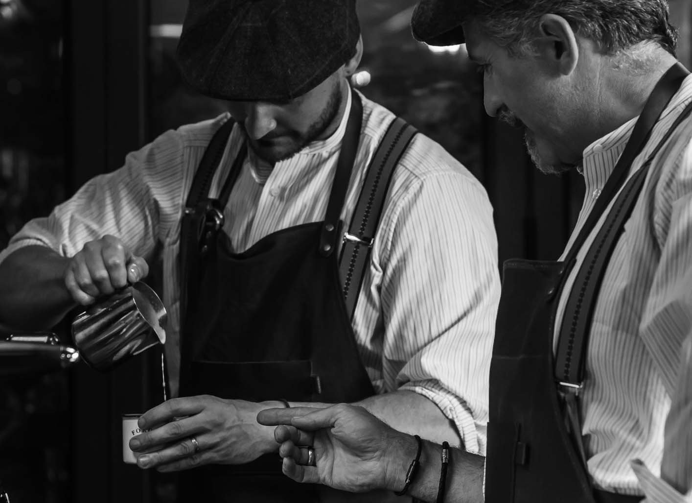 Two men in striped shirts and aprons working together, one pouring coffee into a cup, in a black and white image.
