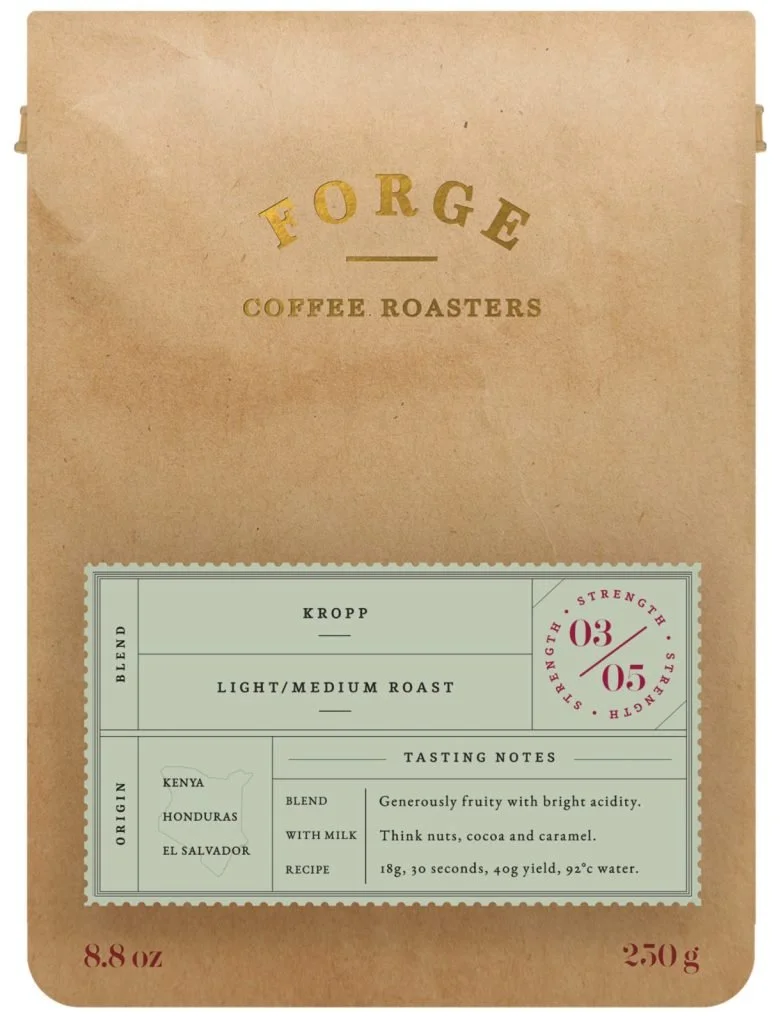 Brown paper bag with gold and black text reading "Forge Coffee Roasters" and a vintage-style coffee label with details about a light to medium roast coffee from Honduras, Kenya, and El Salvador, including tasting notes and brewing instructions.