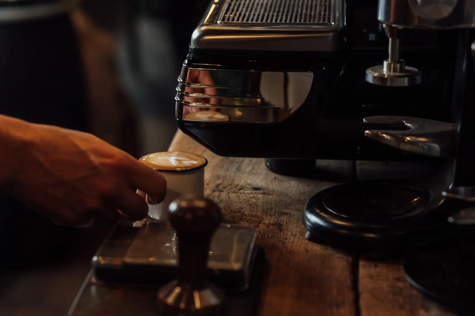 A barista pours steamed milk into a cup of espresso, creating latte art, with a coffee machine on a wooden counter.