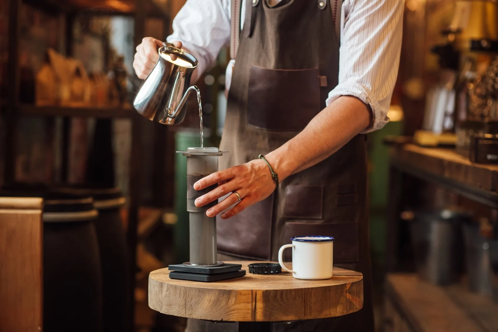 A person in a brown apron pouring hot water from a metallic kettle into a transitional coffee brewing device on a round wooden table, with a white mug nearby, in a cozy wooden cafe setting.