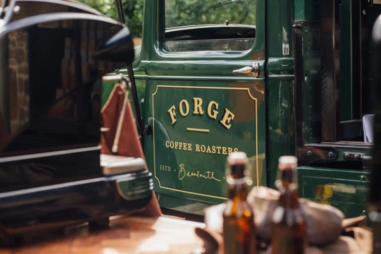 Close-up of vintage green Ford truck with 'Forge Coffee Roasters' logo on the door, in front of a black vintage vehicle, with bottles and items on a wooden surface in the foreground.