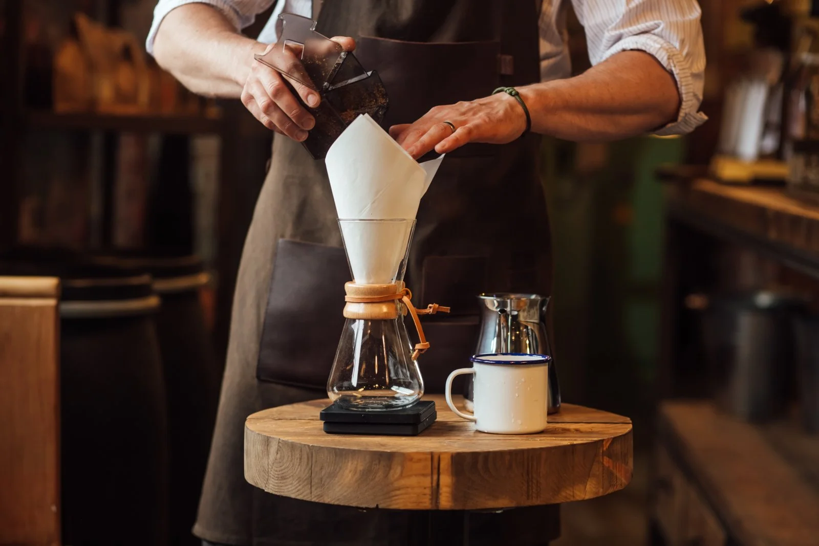 A barista filters coffee into a glass cup through a paper filter held in a funnel over a wooden table, with a mug and carafe nearby.