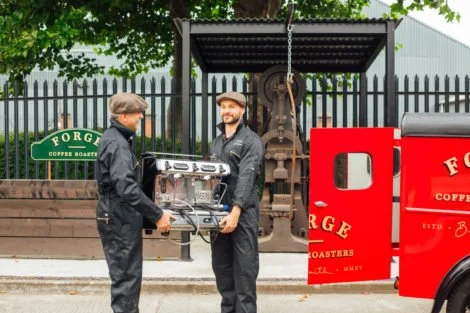Two men in black uniforms shake hands in front of a cafe with a green sign reading "Forged Coffee Roasters." They are standing next to a red delivery truck and a black metal structure with an industrial-like gear. One man is holding a coffee machine.