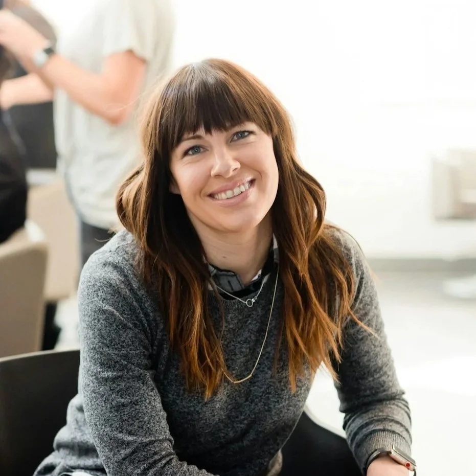 A smiling woman with wavy brown hair and bangs sitting indoors, wearing a gray sweater and a necklace.