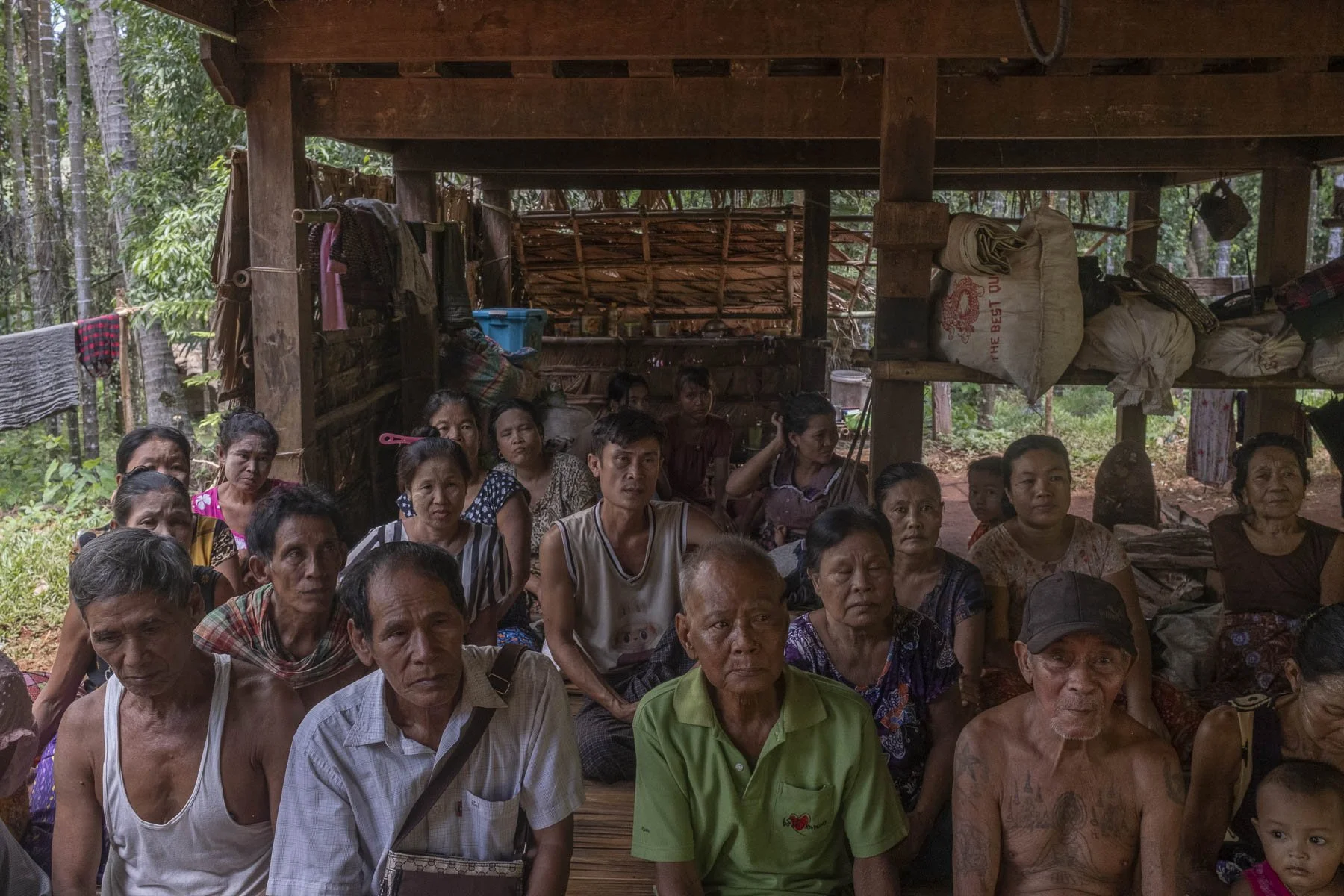 Group of people sitting inside a wooden shelter in a forested area, some wearing casual clothes, with greenery outside.