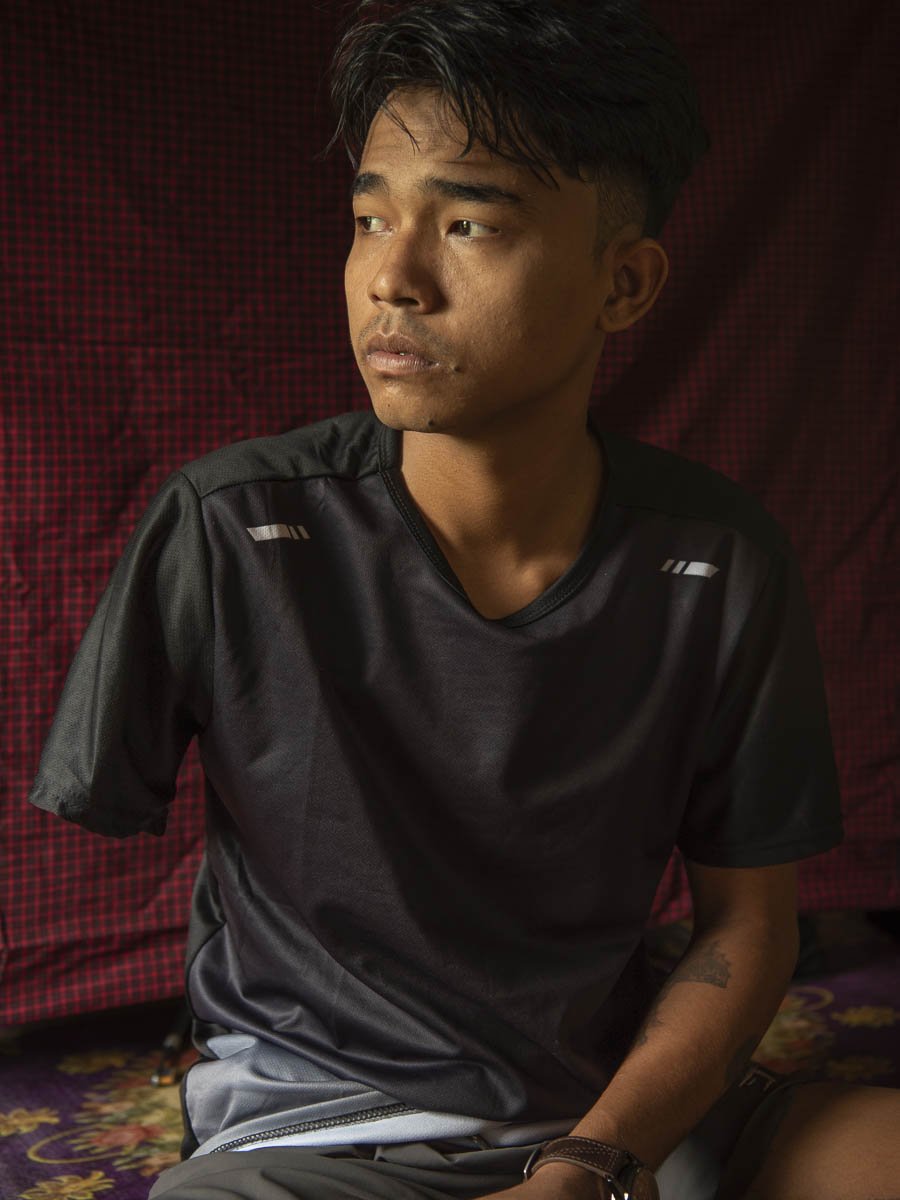 A young man with short dark hair and a tattoo on his left forearm sitting against a red and black checkered background, wearing a black T-shirt.