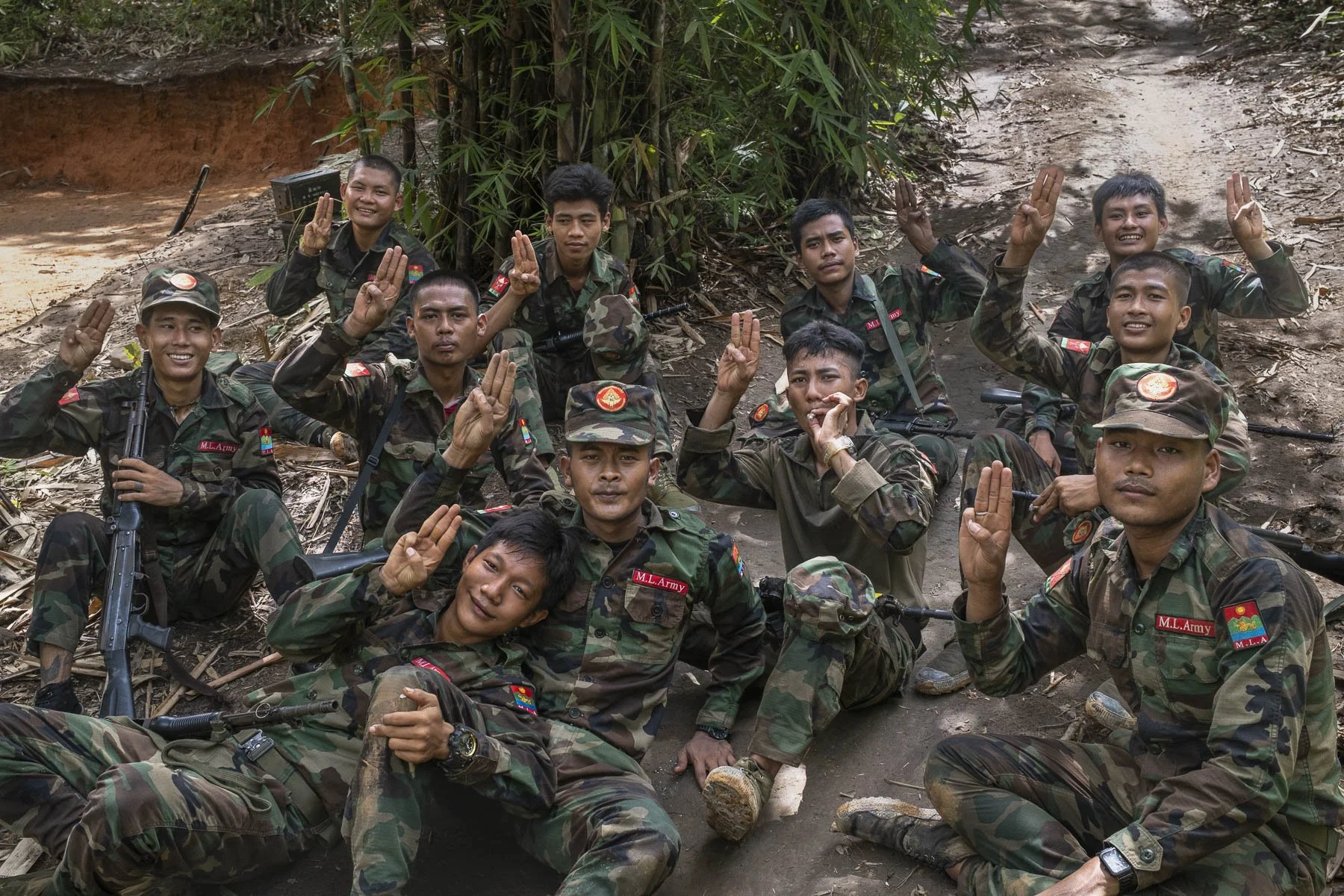 A group of soldiers in camouflage uniforms sitting and kneeling outdoors, making the three-finger salute, with trees and dirt trail in the background.
