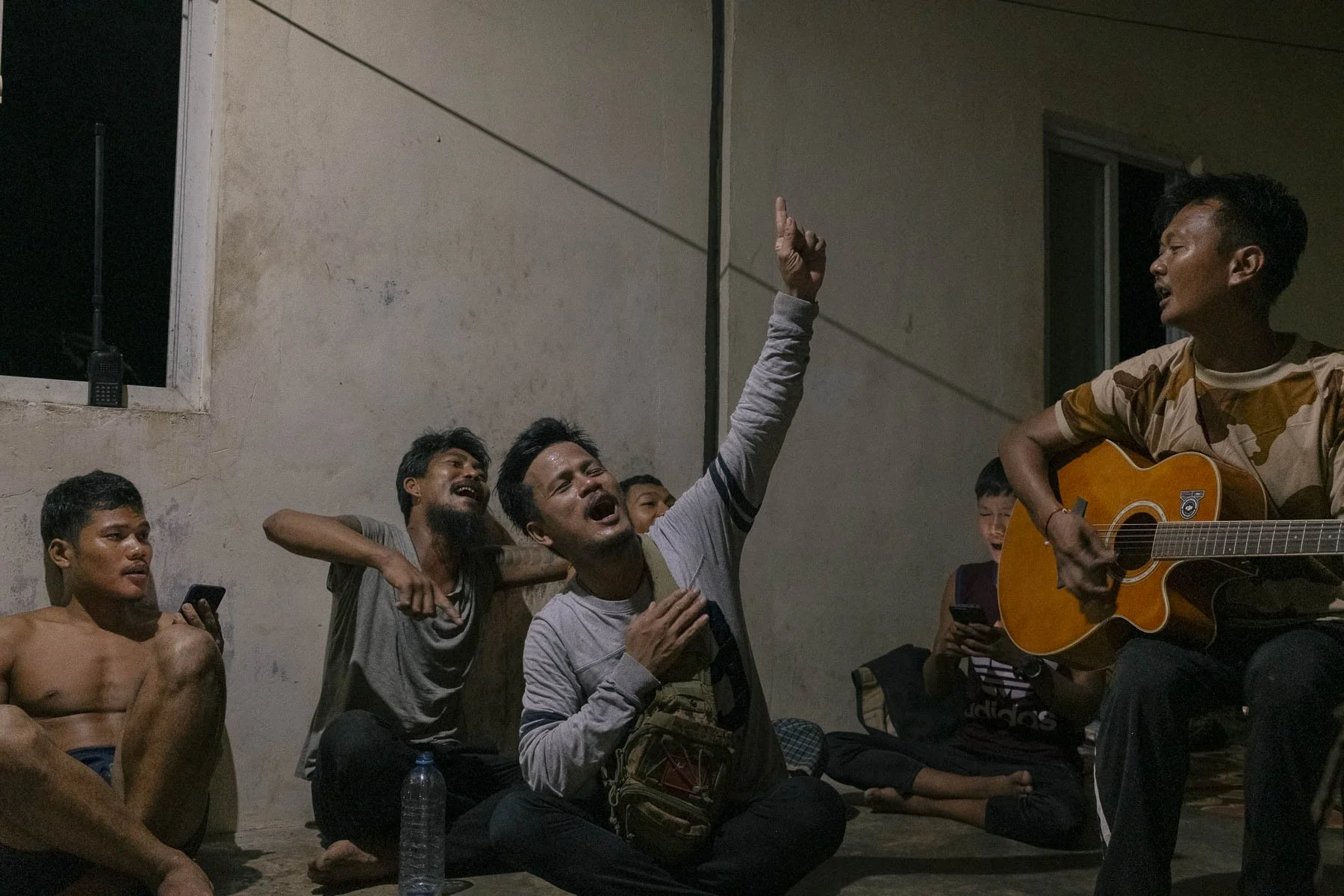 A group of young men sitting in a room, with one playing a guitar and others singing along passionately, some using phones, during an indoor musical gathering at night.