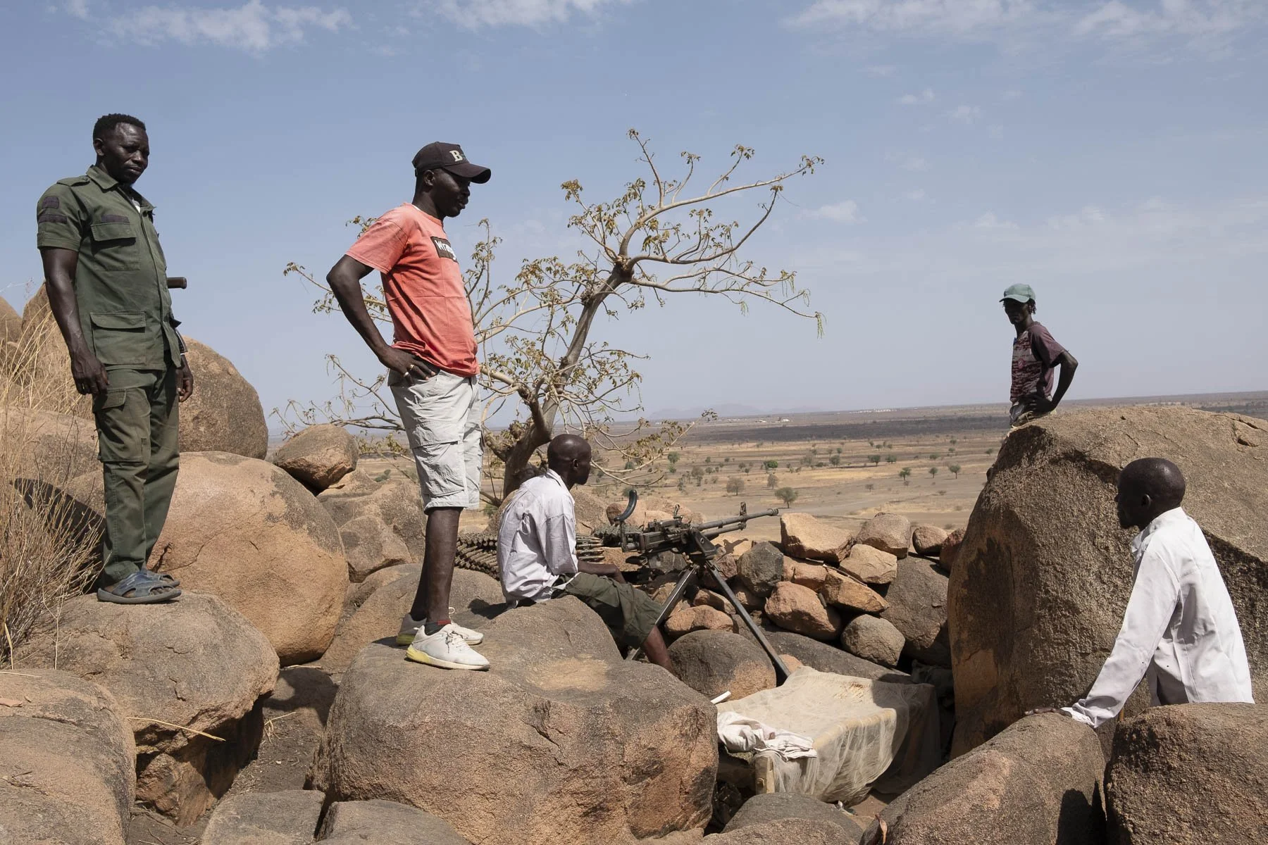Five men in a rocky landscape with a tree and open land in the background. One man is standing on the left, dressed in a green military-style uniform. Two men are standing in the center, one wearing a red t-shirt and cap, and the other sitting on a r