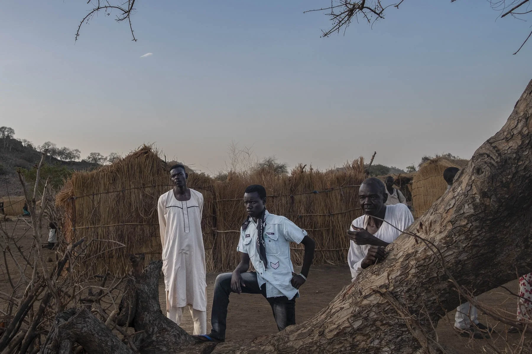 Three men standing outside near a large fallen tree and straw huts, with a clear sky and distant hills in the background.