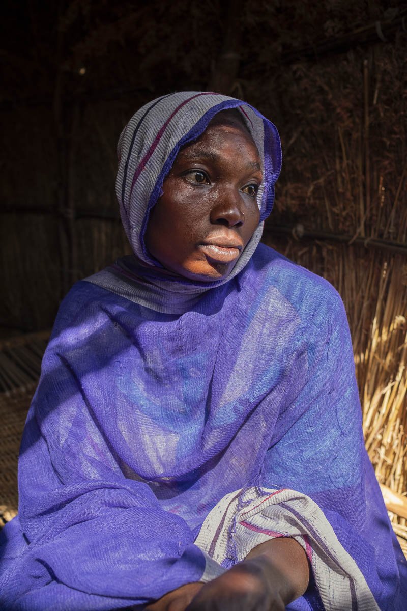 A woman wearing traditional clothing and a headscarf, sitting inside a hut made of natural materials. She appears to be deep in thought.