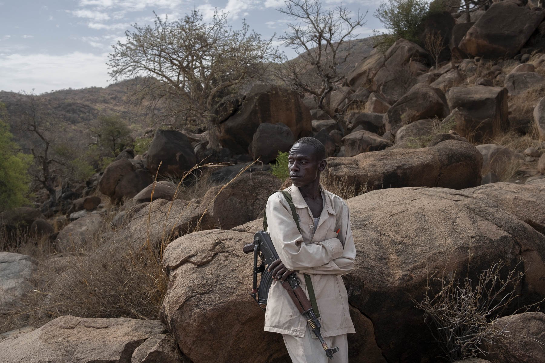 A man in a beige outfit standing among rocks in a dry, mountainous landscape, carrying a rifle over his shoulder.