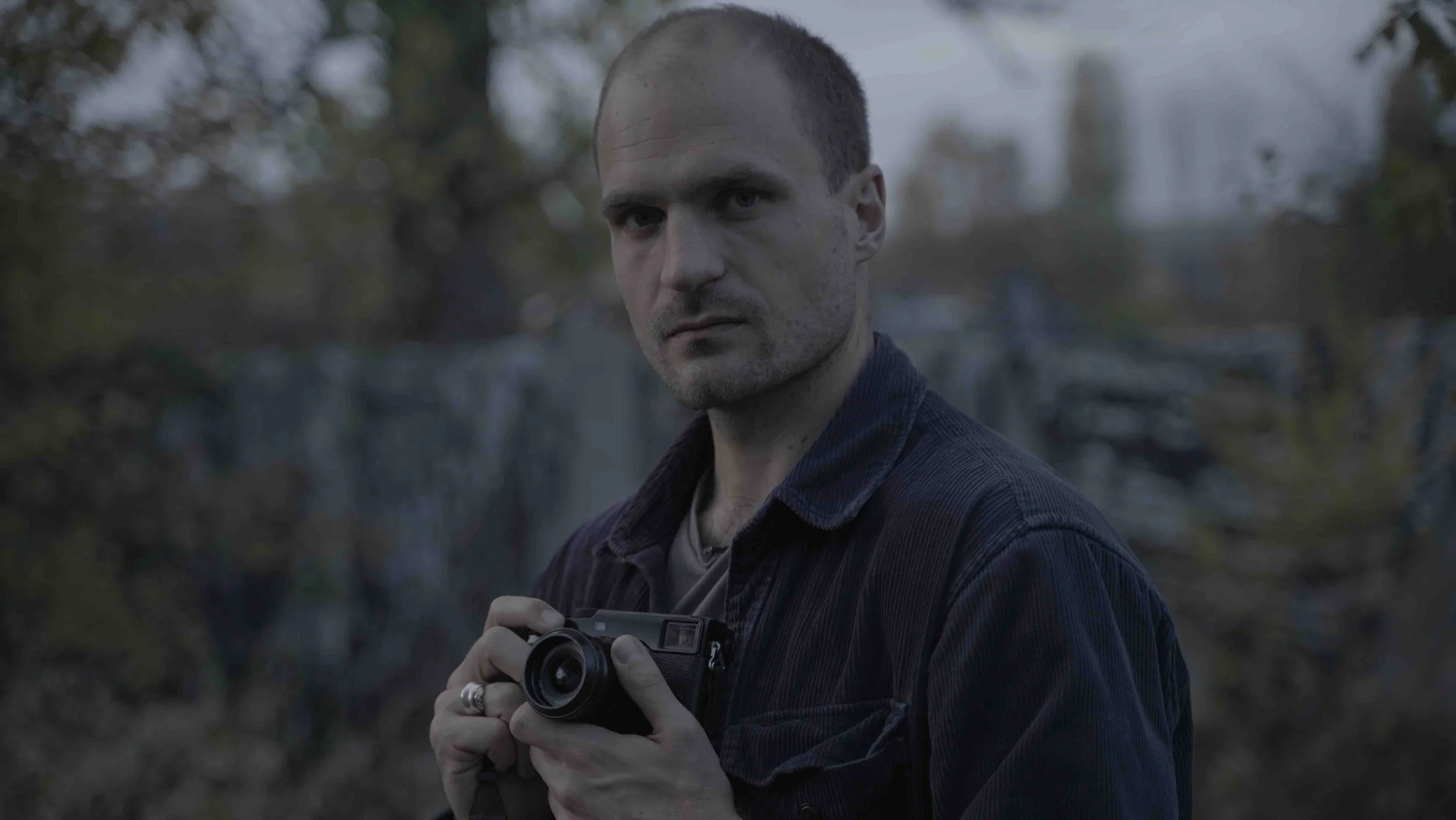 A man with short hair and a serious expression holding a vintage camera outdoors near a waterfall, with trees and rocks in the background during dusk or early evening.