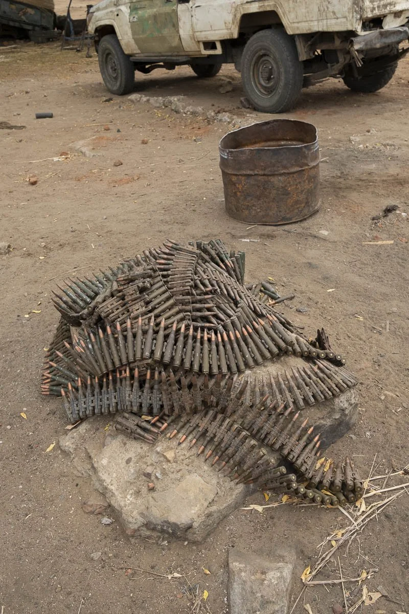 A pile of bullets arranged to resemble a crocodile, placed on a large rock in front of a rusty barrel and an off-road vehicle in a barren outdoor setting.