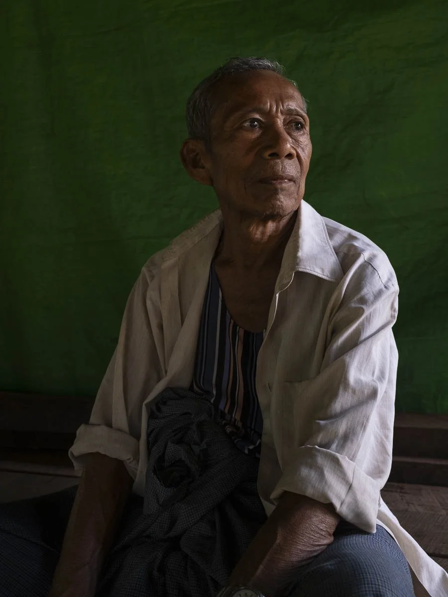 An elderly man with gray hair and a thoughtful expression, wearing a light-colored shirt over a striped undershirt, sitting against a dark green background.