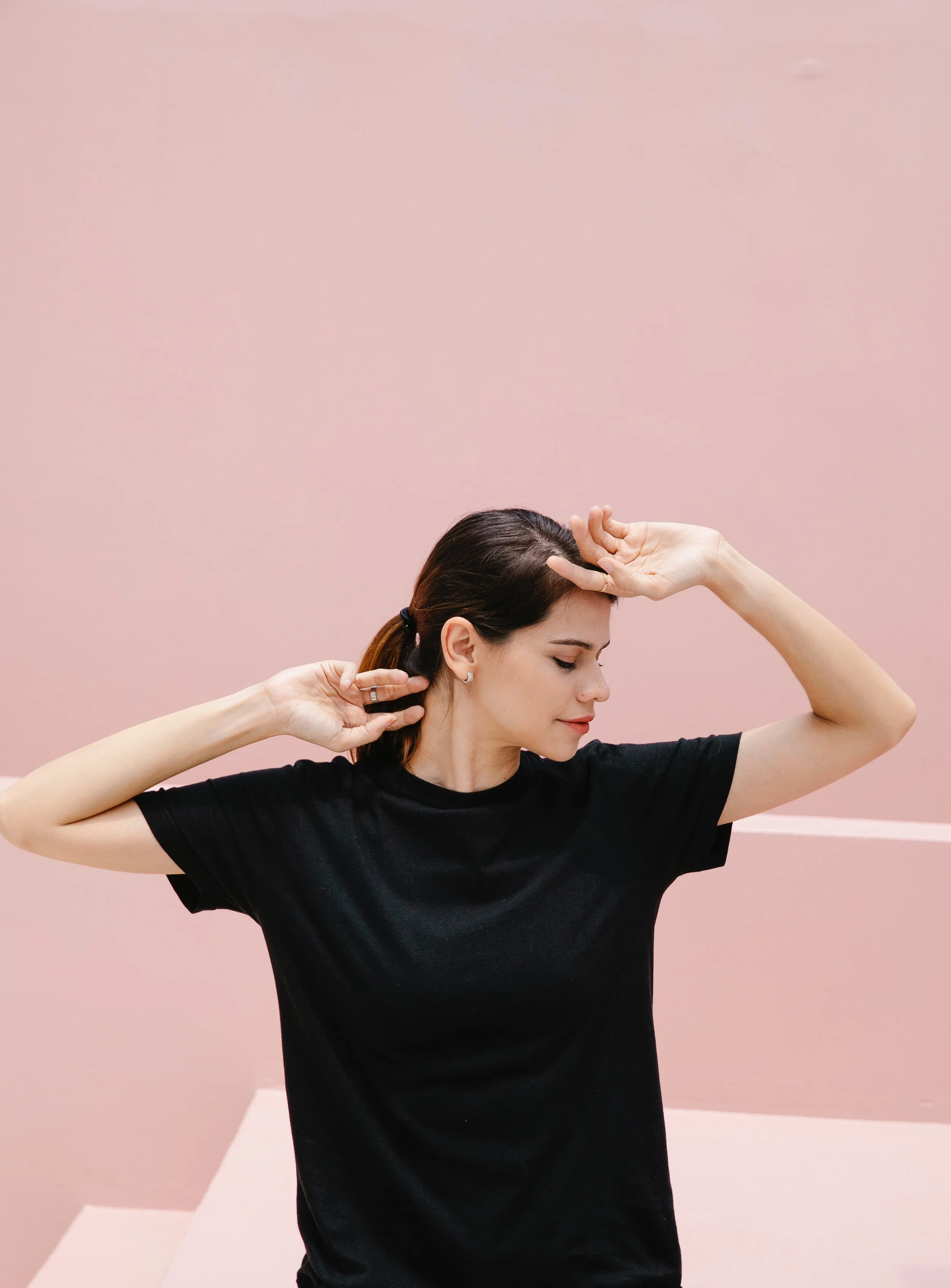 Young woman with brown hair in a ponytail, wearing a black t-shirt, standing against a pink background, with her eyes closed and hands on her forehead and neck, appearing relaxed or stretching.