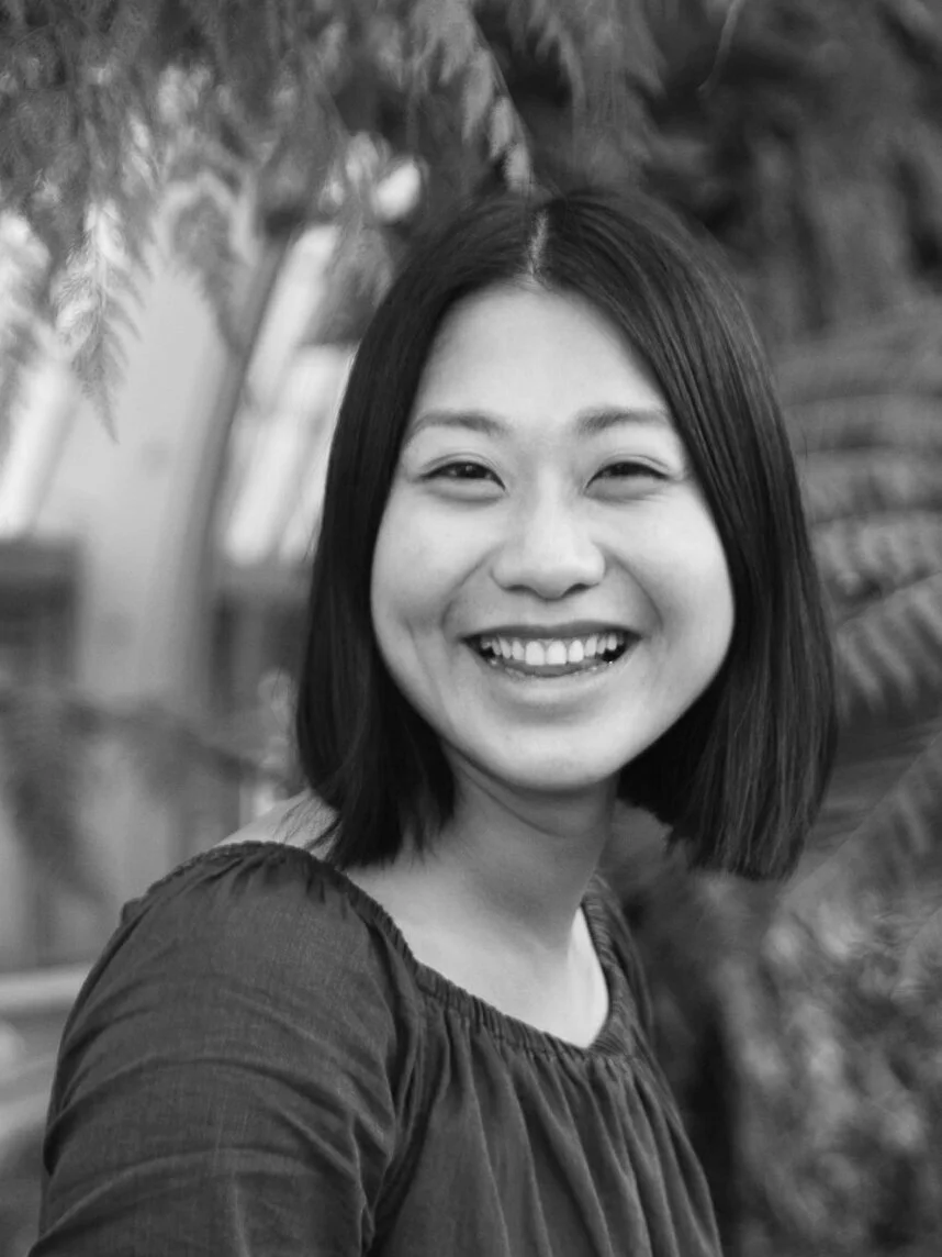 Black and white photo of a woman with shoulder-length dark hair smiling outdoors, surrounded by plants.
