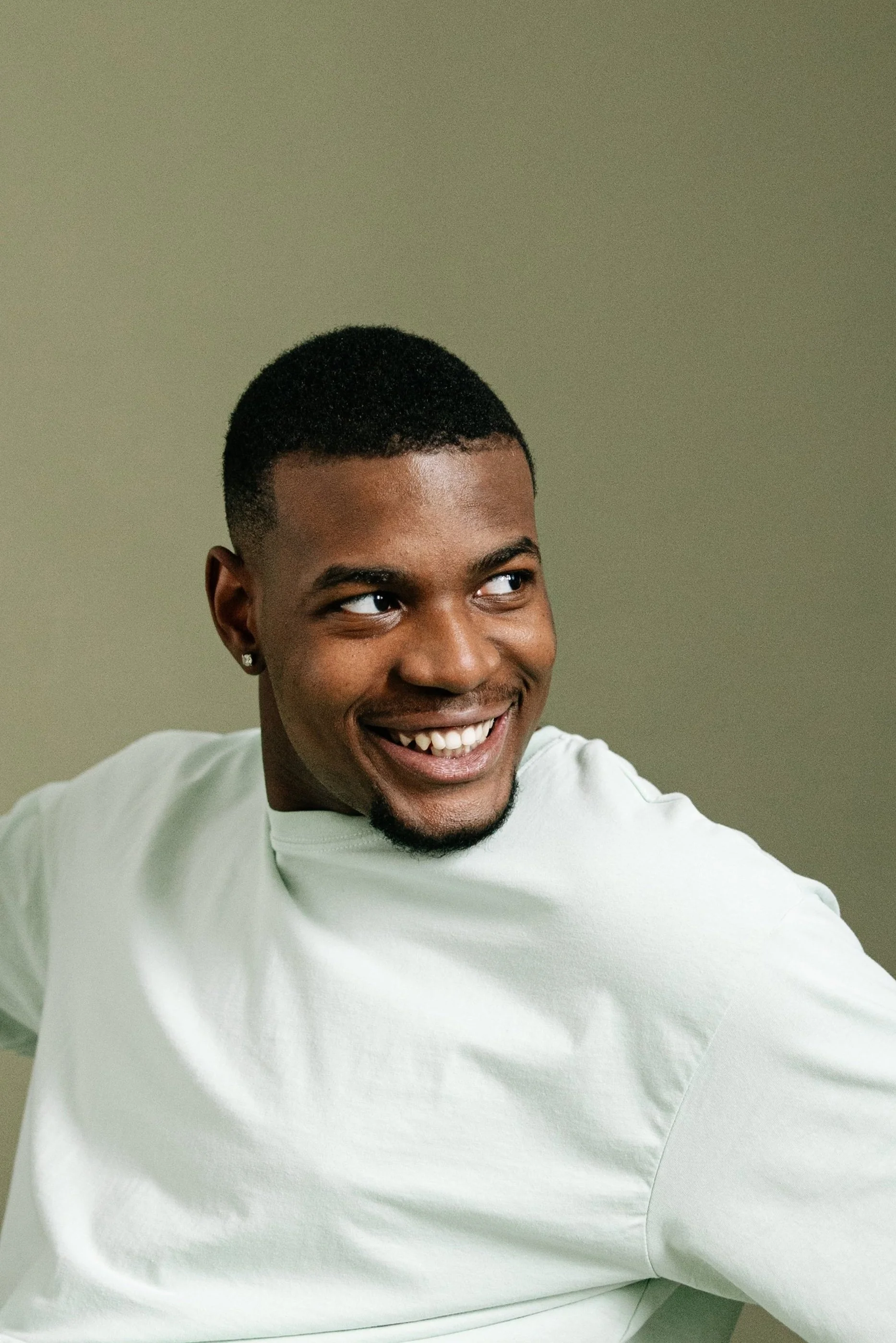 A young man with a short haircut and earrings, smiling and looking to his right, wearing a white shirt, against a plain background.
