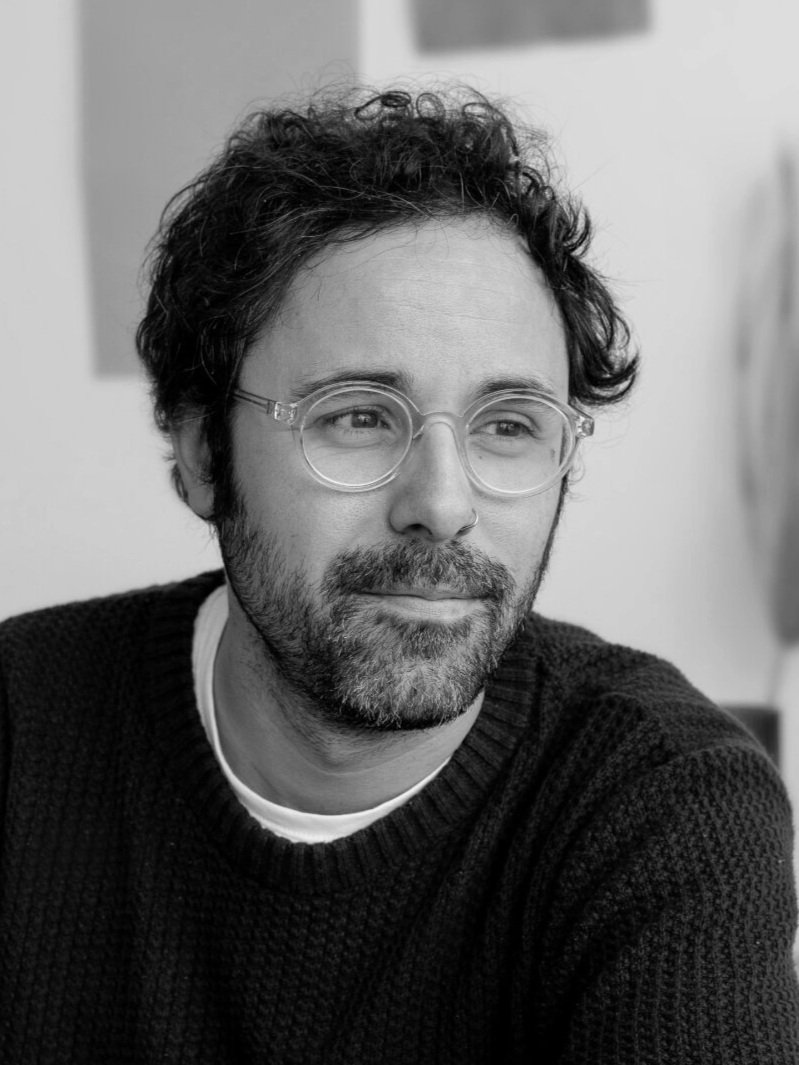 A man with curly hair, glasses, and a beard, wearing a black sweater and a white shirt underneath, looking thoughtfully to the side in a black and white photo.