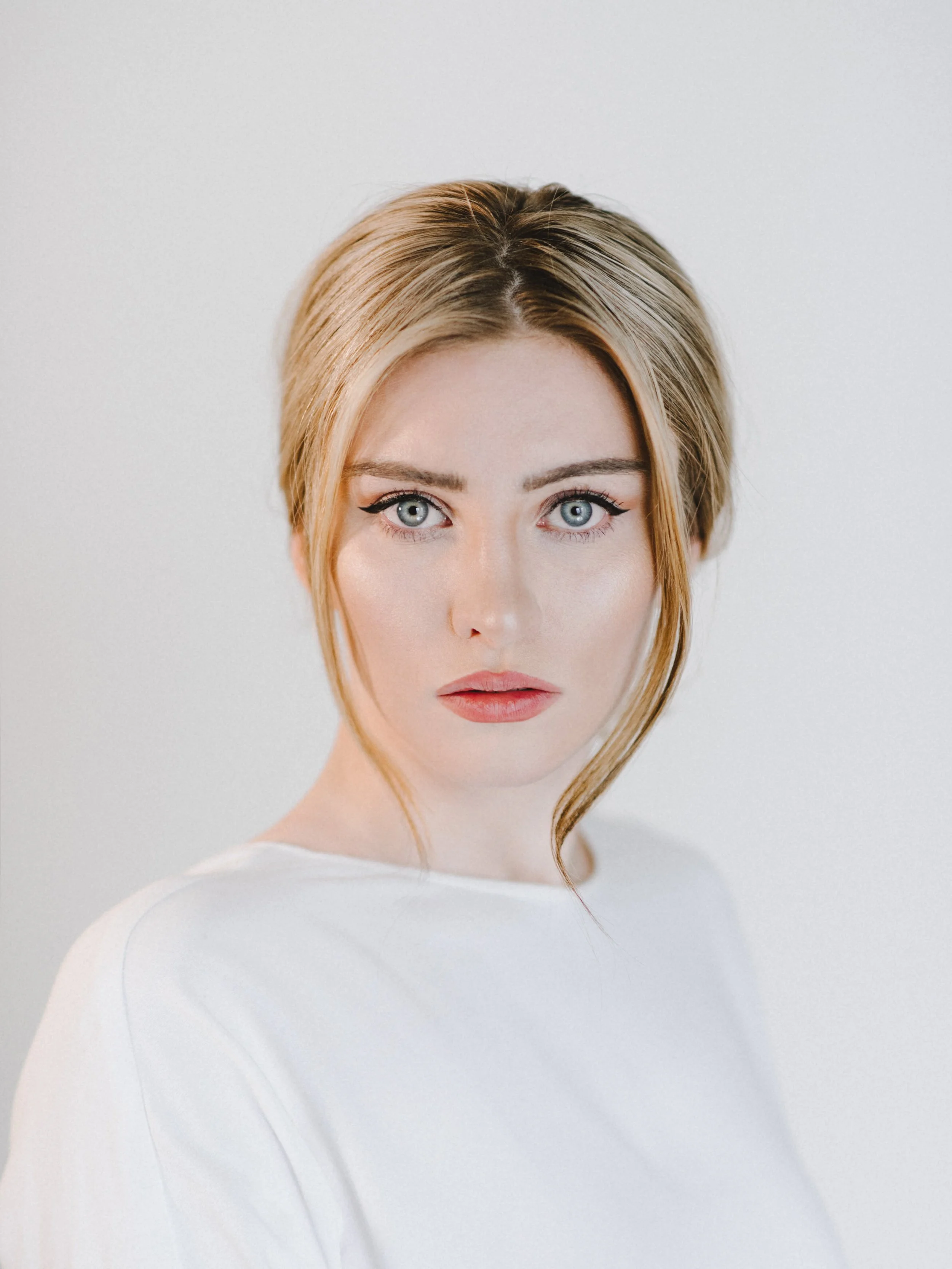 A portrait of a young woman with blonde hair and blue eyes, wearing a white top, looking directly at the camera with a neutral expression.