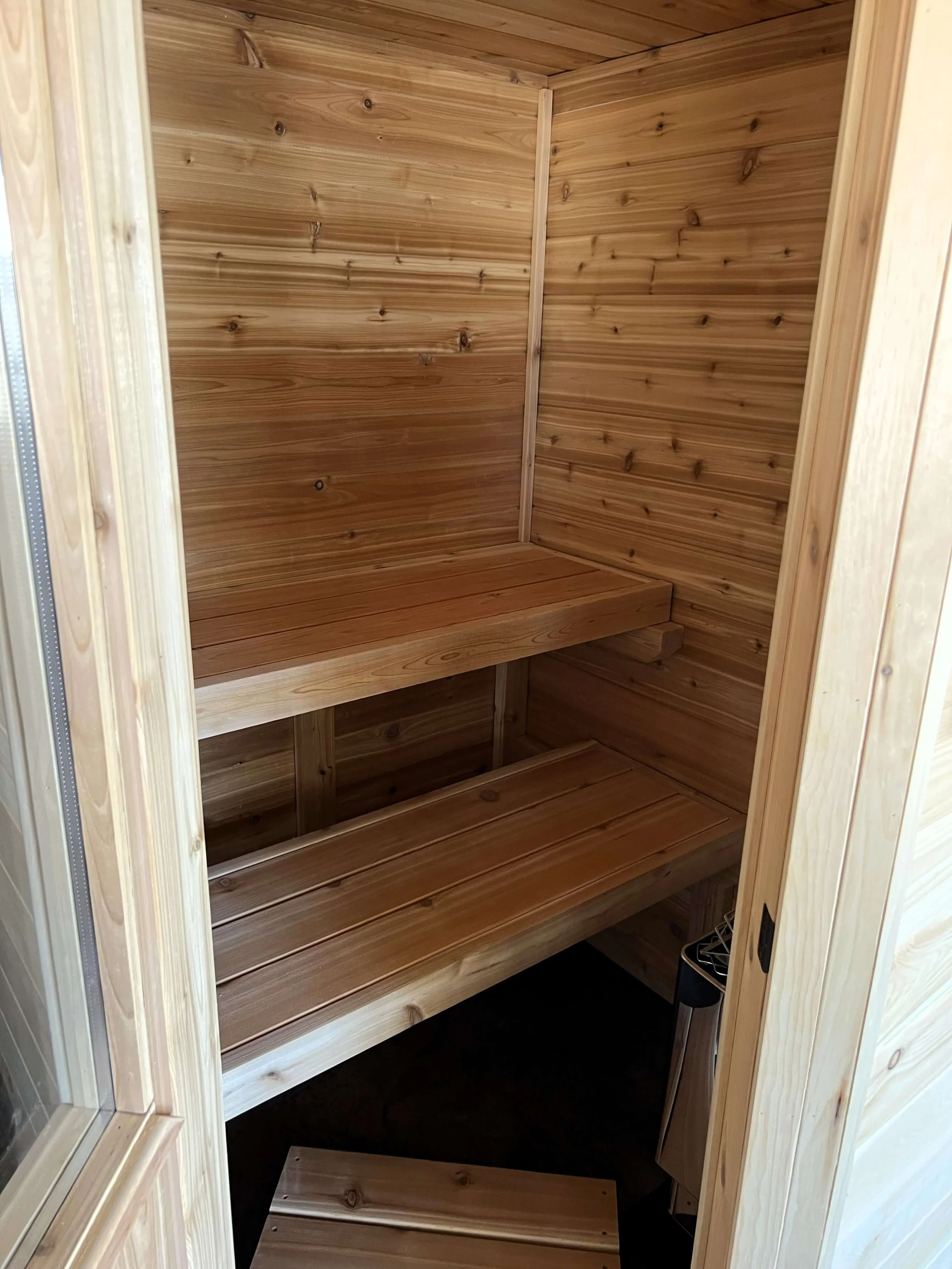 Open door view of custom Western Red Cedar electric sauna interior showing upper and lower benches with natural lighting.