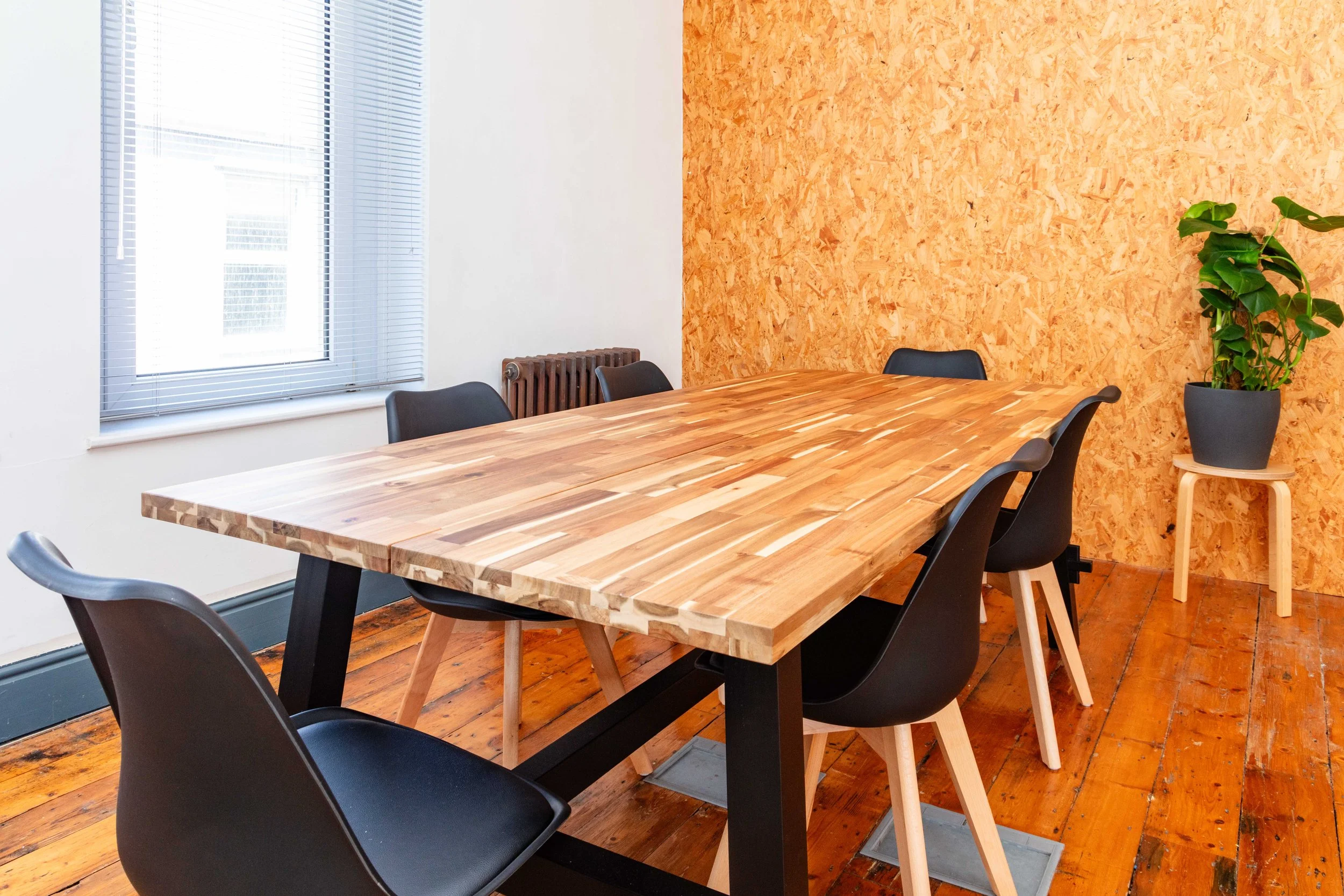 A meeting room with a wooden table, black chairs, a plant in a pot, and a window with blinds, featuring a wooden wall and wooden floor.
