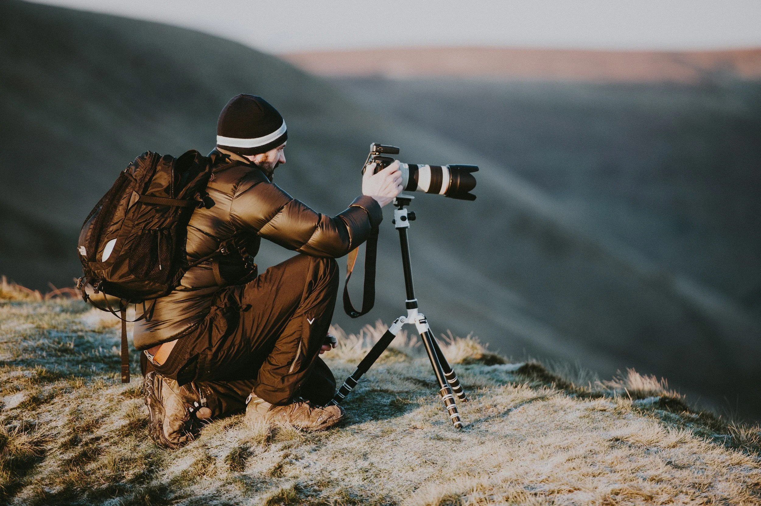 Photographer kneeling on grassy hill, adjusting camera on tripod, overlooking mountainous landscape during sunset.