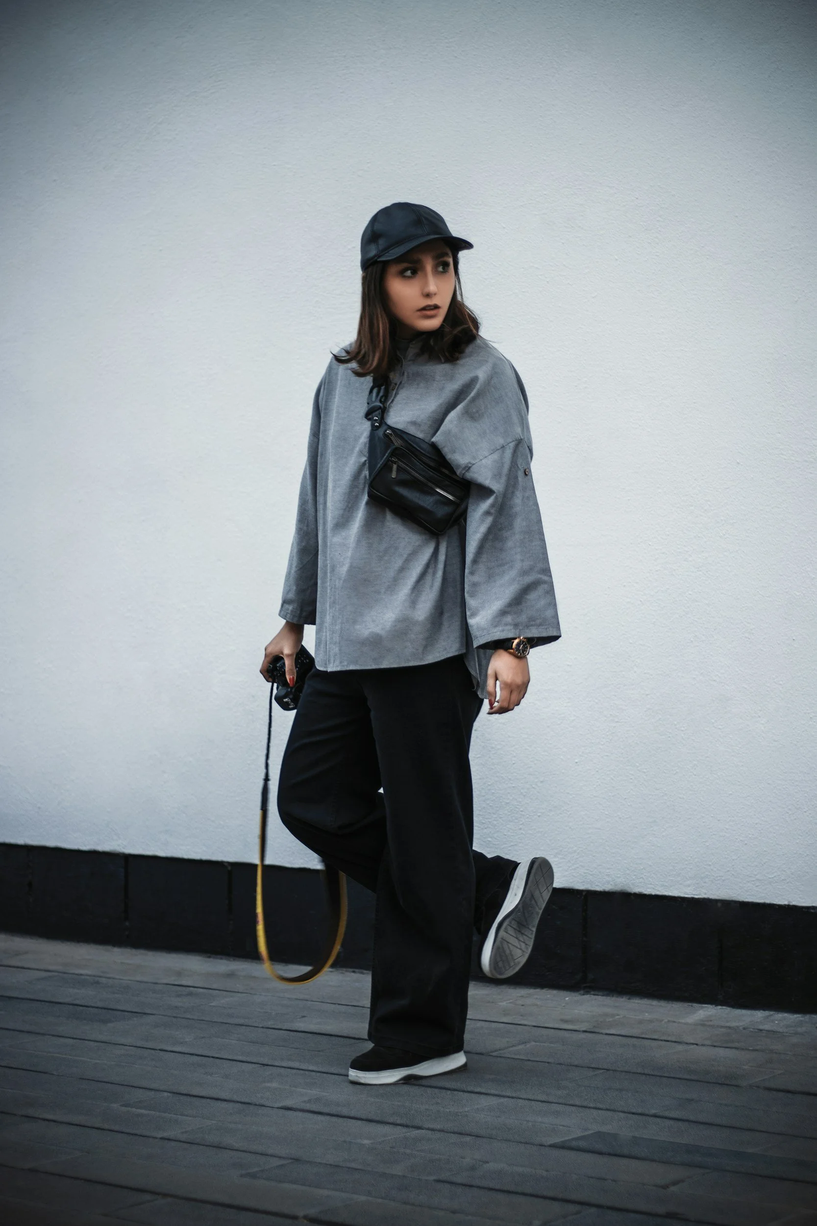A woman dressed in casual, dark-colored clothing standing outdoors against a white wall. She is wearing a black cap, a gray oversized jacket, black pants, and sneakers. She is holding an object in her right hand and has a small black pouch around her chest.