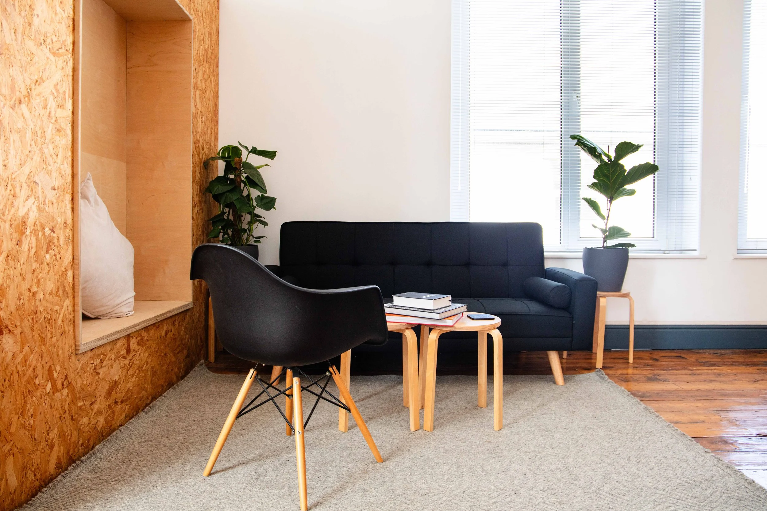 Modern living room with black sofa, black armchair, small wooden coffee table with books, potted plants, large windows with blinds, and a beige carpet on wooden floor.