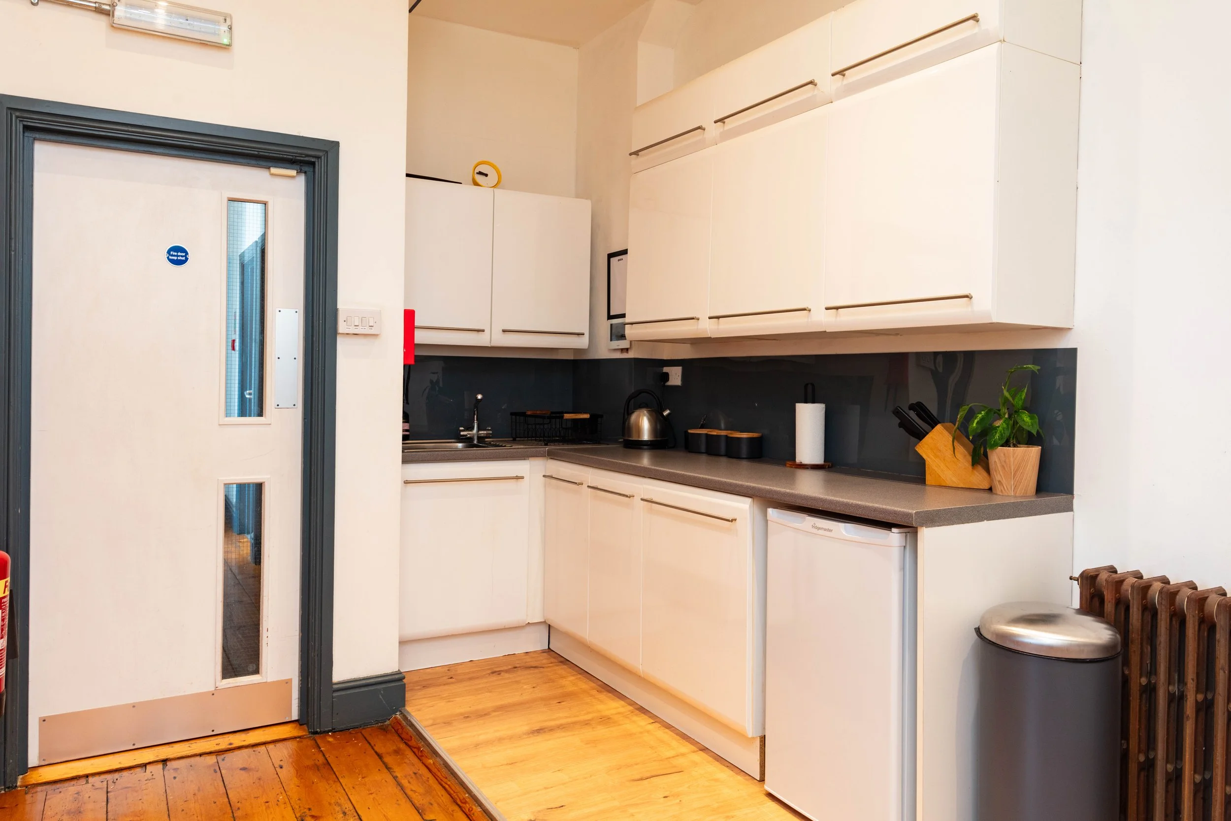 A small kitchen with white cabinets, a gray countertop, a tiny refrigerator, a trash can, and a door with a small window.