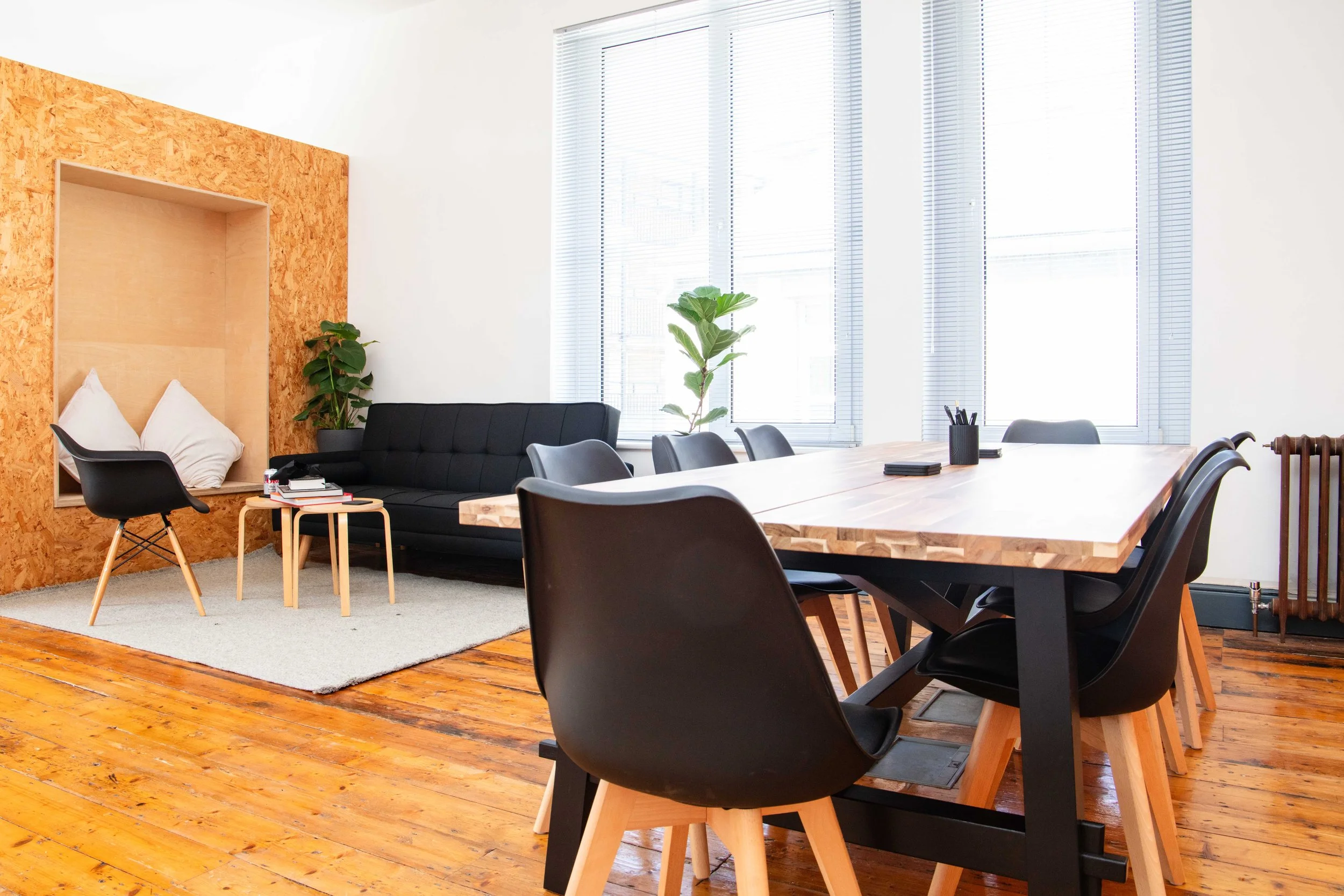 Modern conference room with large wooden table, black chairs, and a cozy seating area with black sofa, small side tables, and a cushioned nook with pillows. Bright natural light from large windows with blinds, wooden flooring, and indoor plants.