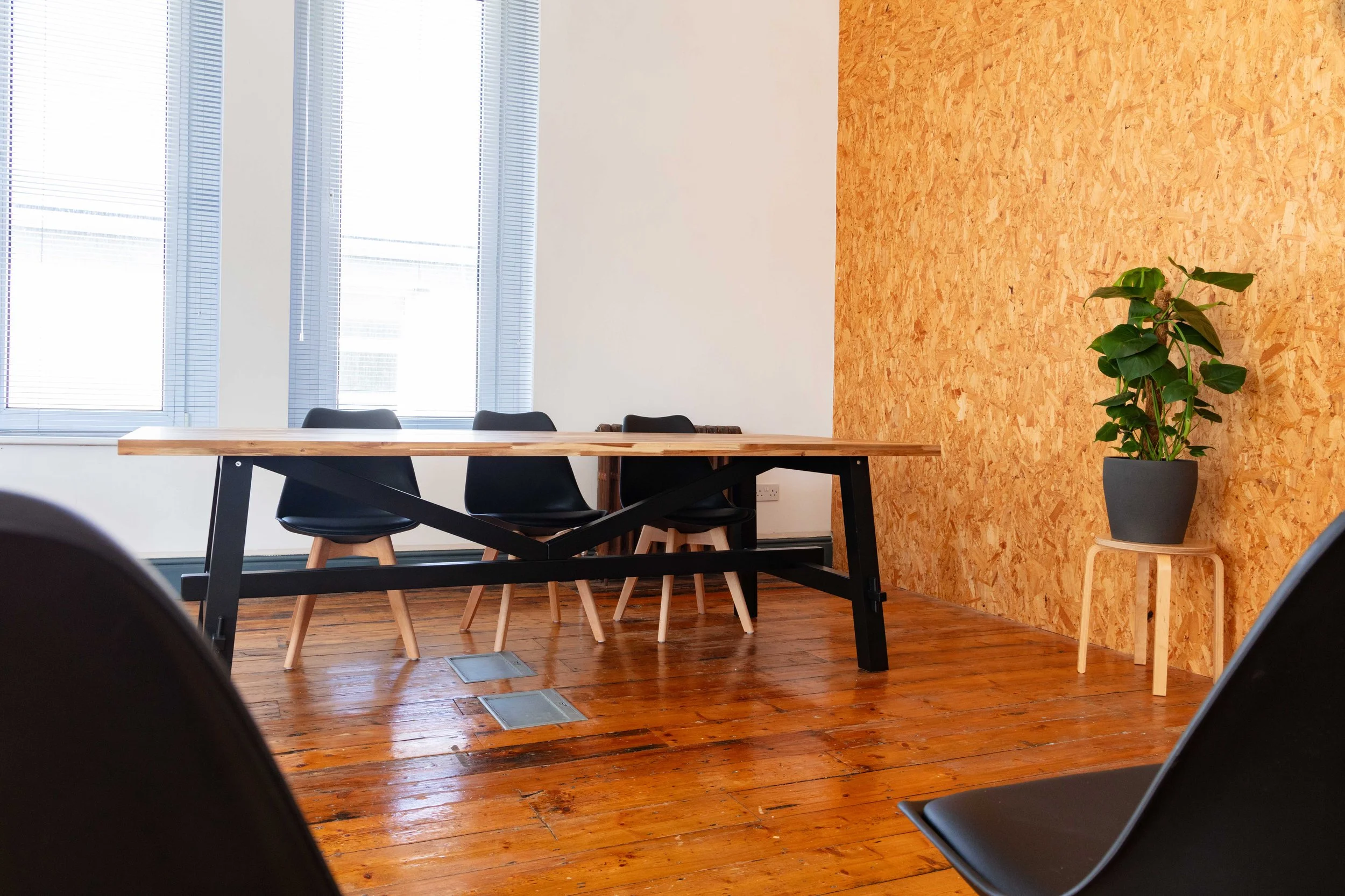 A modern conference room with wooden floor and a large wooden table, black chairs, large windows with blinds, a potted plant on a small table, and an orange OSB wall.