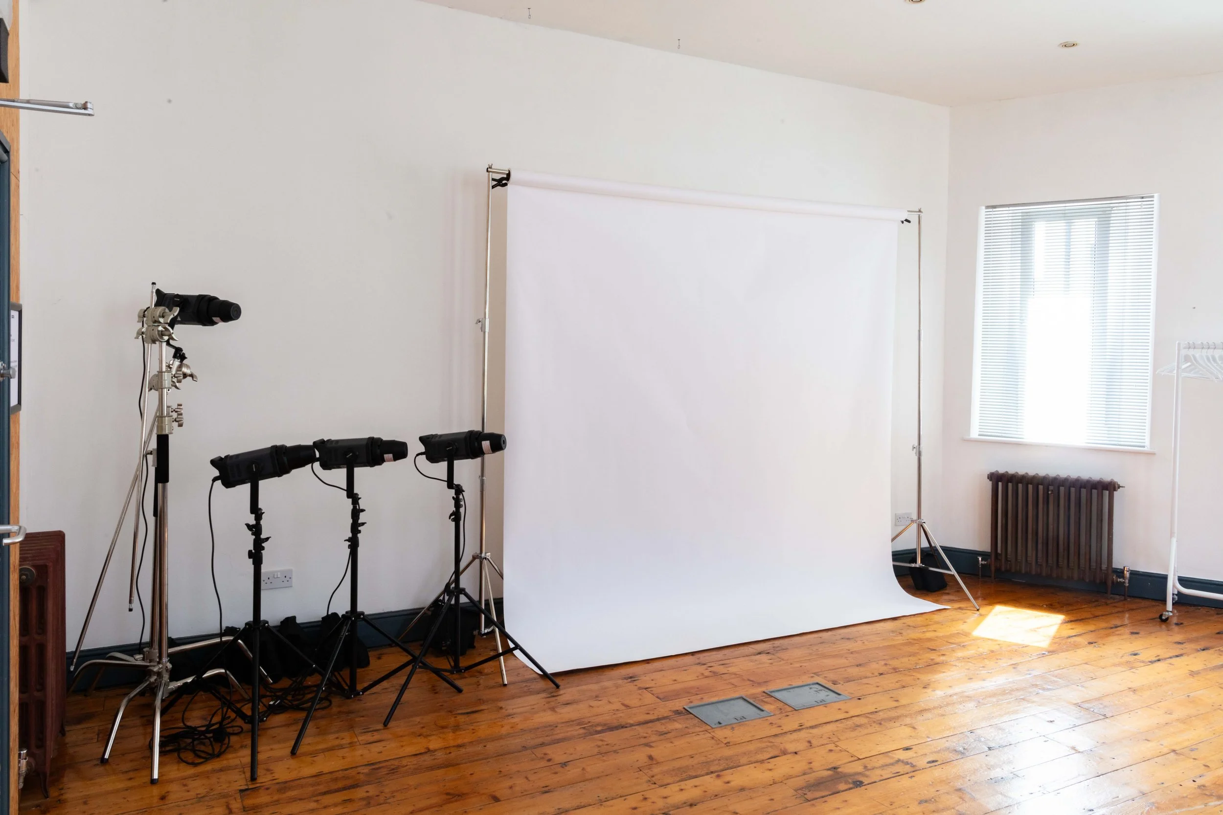 Photography studio setup with a white backdrop, three studio lights on stands, a window with blinds, and a radiator below the window.
