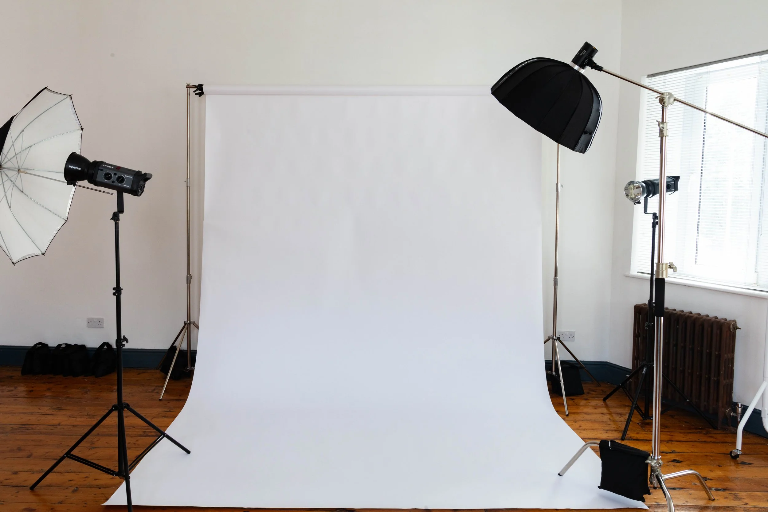 Photography studio setup with a white backdrop, two softbox lights, two umbrella lights, and camera accessories on the floor.
