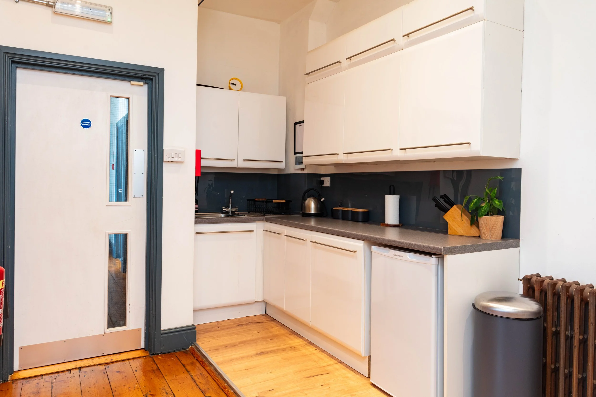 Small kitchen with white cabinets, a gray countertop, and a small fridge. On the countertop, there is a paper towel holder, a potted plant, and a wooden knife block. A metallic kettle is near the sink. A black dish rack is above the sink, and a blue-