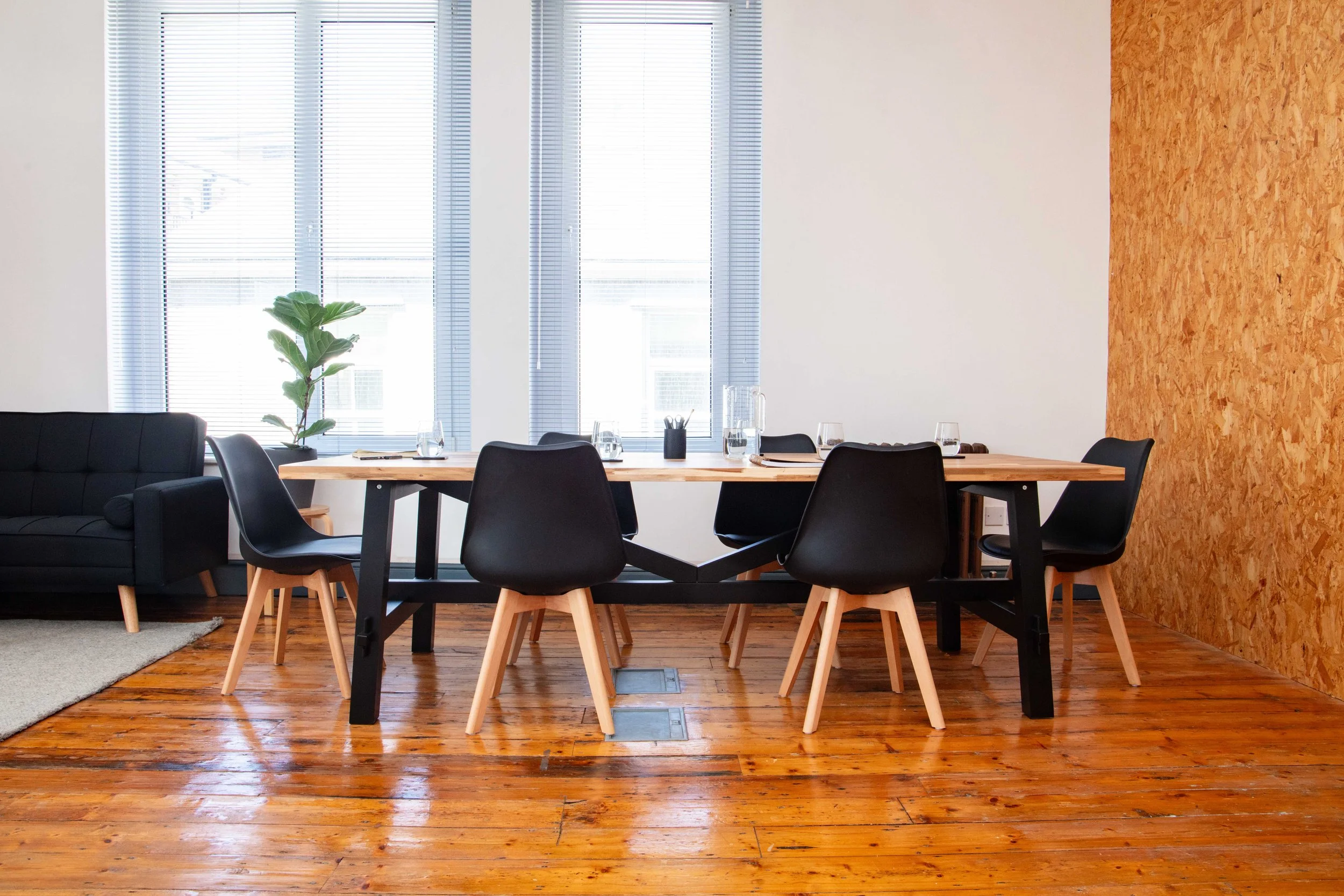 A modern dining room with a wooden table, black chairs with wooden legs, a black sofa, a green potted plant, wooden floors, and large windows with blinds.