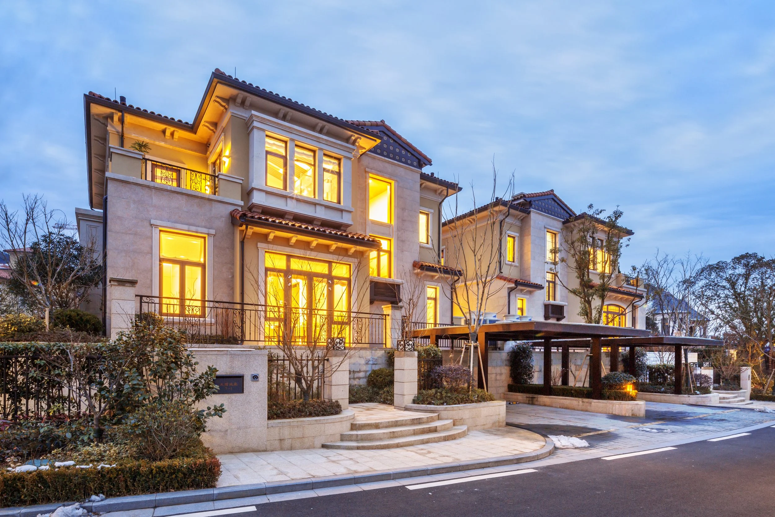 A modern multi-story house with large illuminated windows during dusk, surrounded by trees and a paved driveway with snow patches.