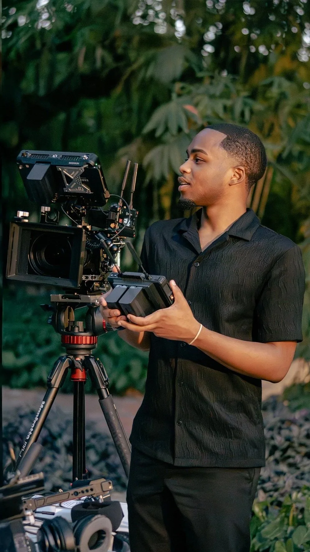 A man operating a professional camera on a tripod outdoors, with lush green foliage in the background.