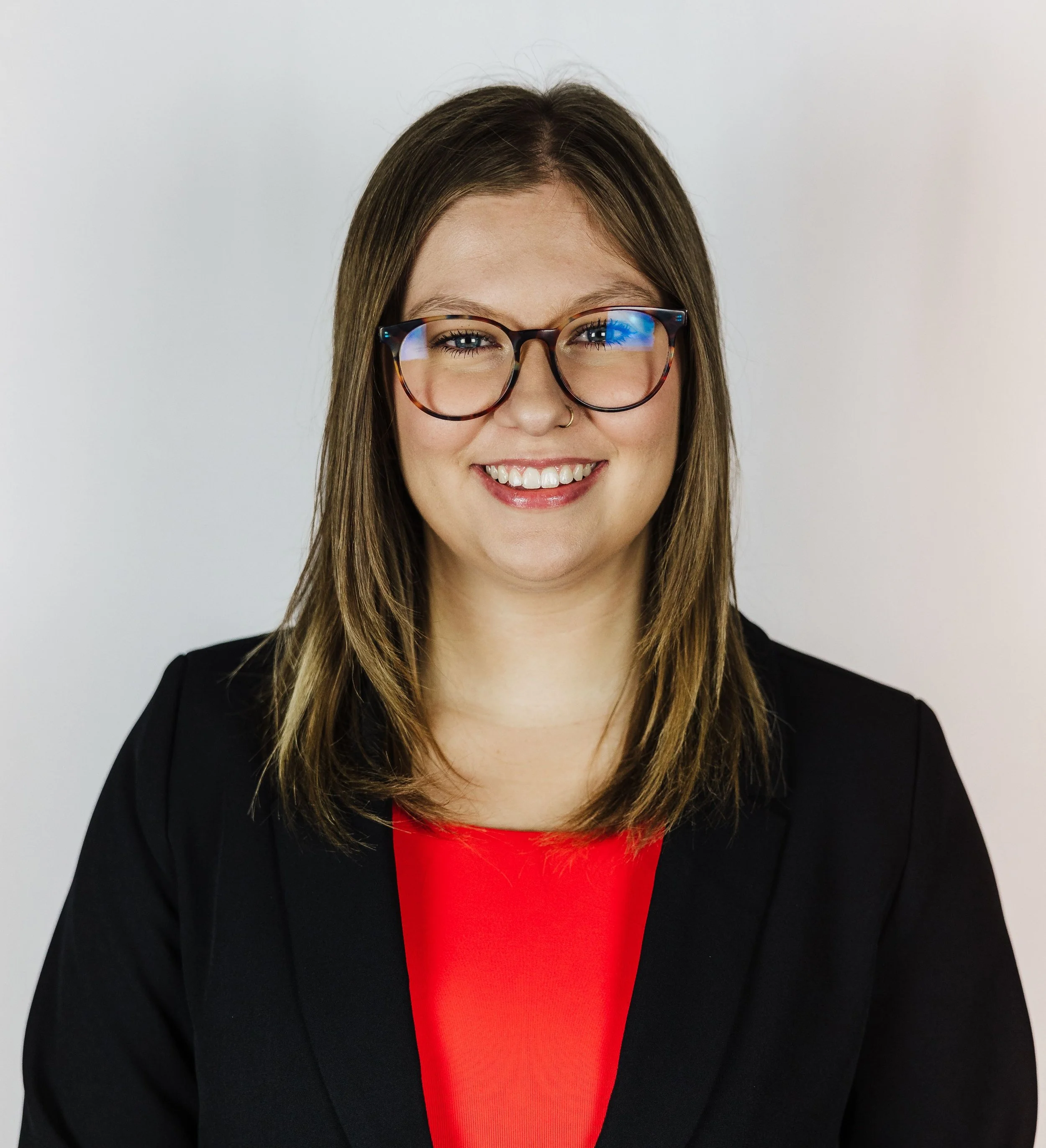 A smiling woman with shoulder-length brown hair, wearing glasses, a black blazer, and a red top against a plain white background.
